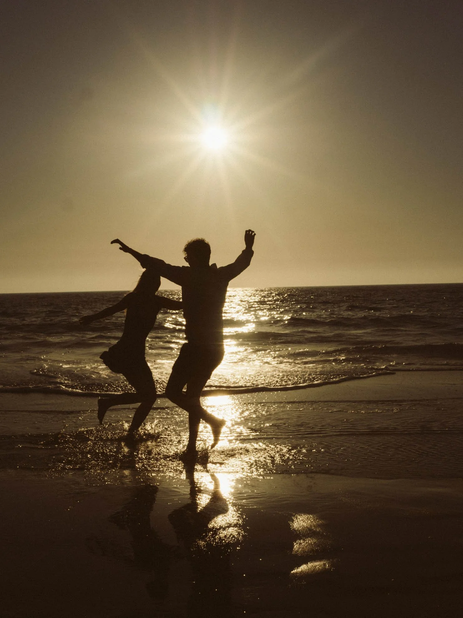Silhouettes of two people playing on the beach at sunset with the sun shining brightly in the sky and reflecting on the water.