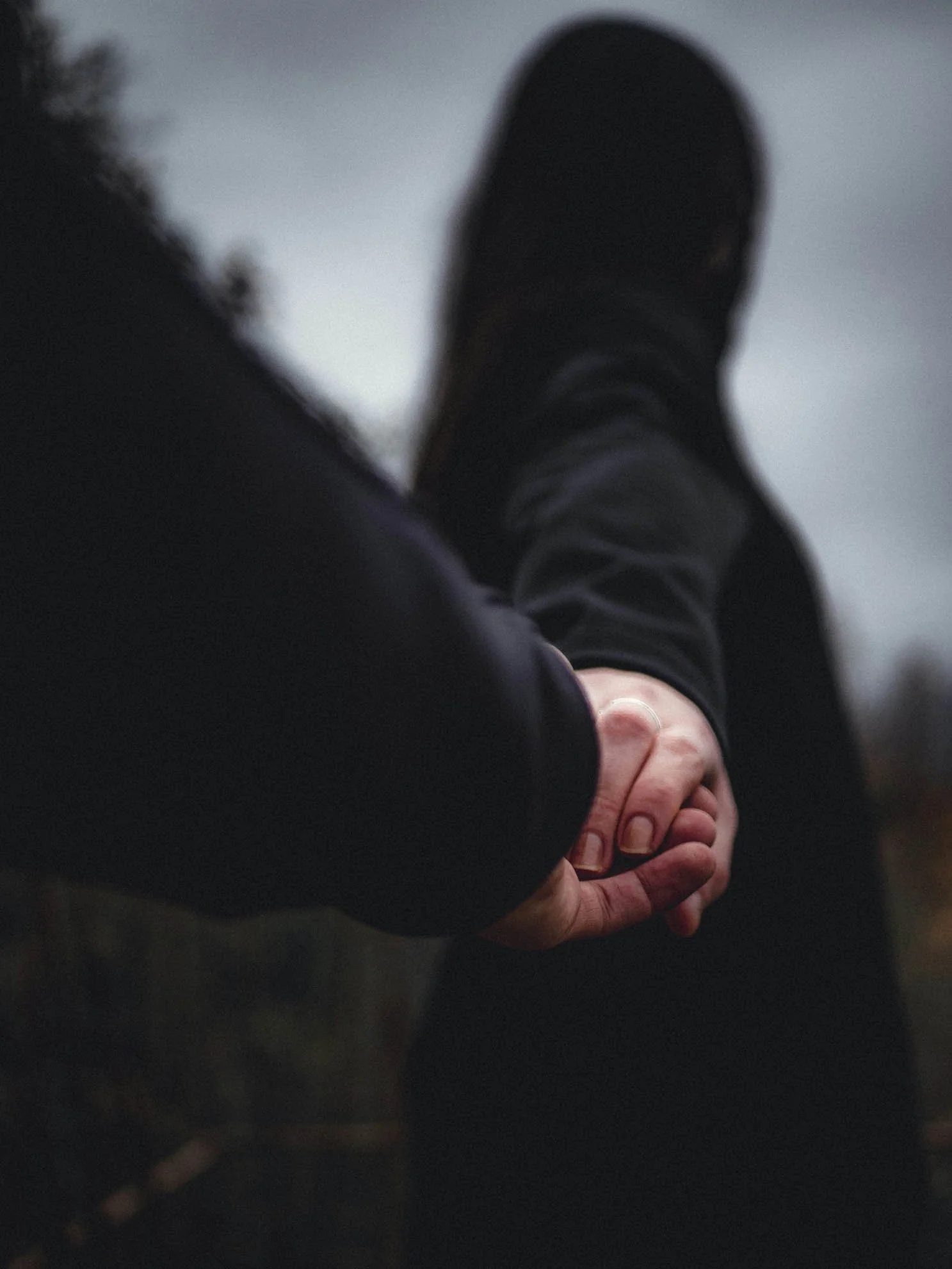 Close-up of a person's hands clasped together resting on legs, with black shoes and dark pants, with a blurred outdoor background.