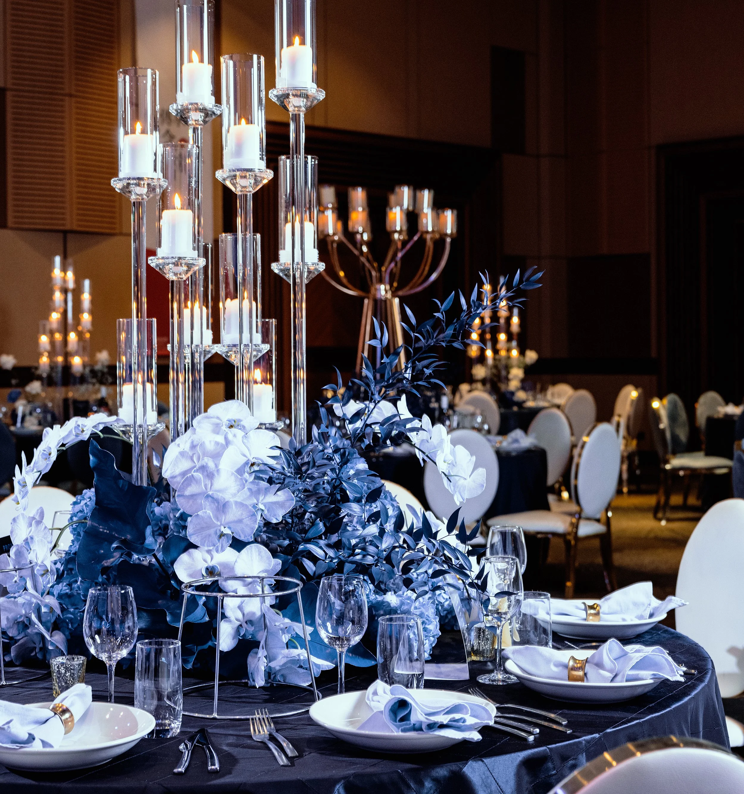 Elegant event table setting with white floral centerpiece and tall candle holders, surrounded by decorated tables and chairs.