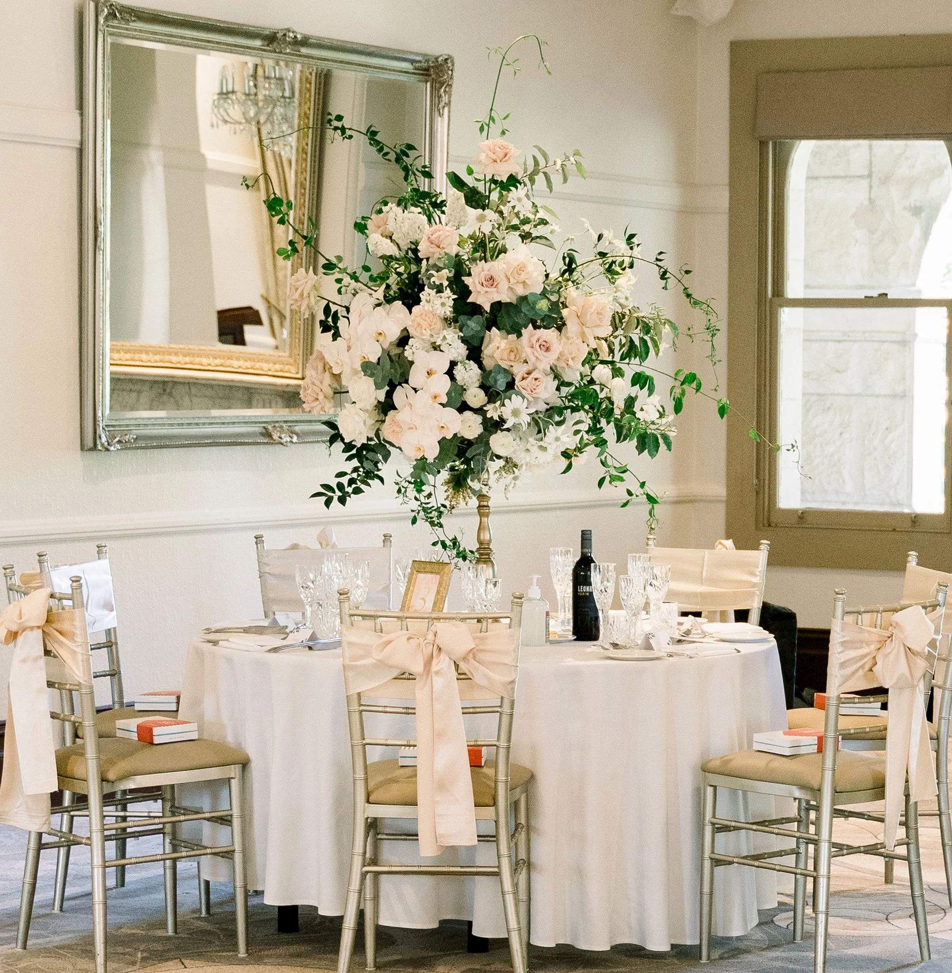Elegant wedding reception table with floral centerpiece, white tablecloth, bow-adorned chairs, and wine bottles in a decorated room with a large mirror and window.