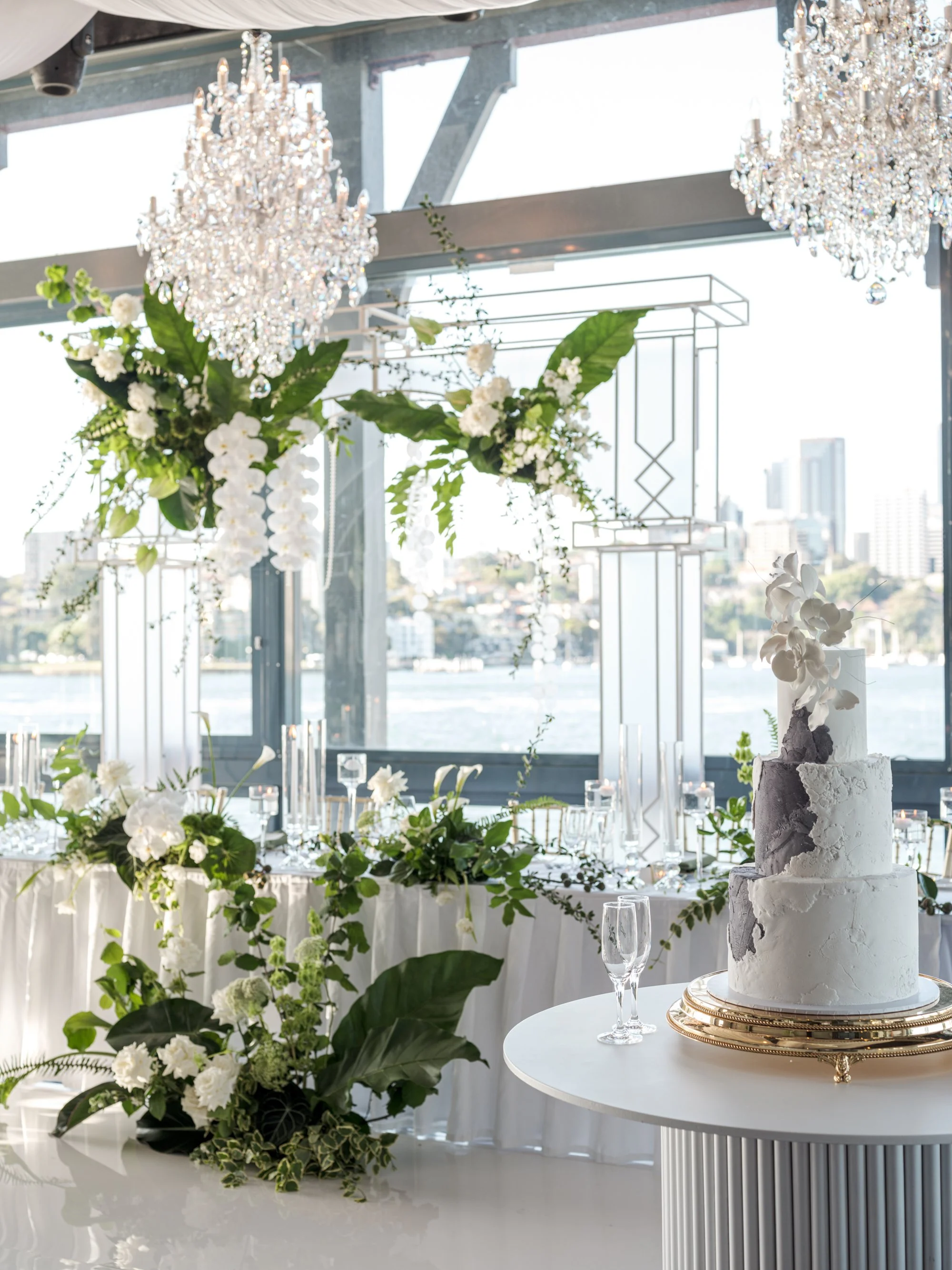Elegant indoor wedding reception with a three-tier white cake on a round table, lush green and white floral arrangements, hanging crystal chandeliers, and a decorated seating area by large windows overlooking water.