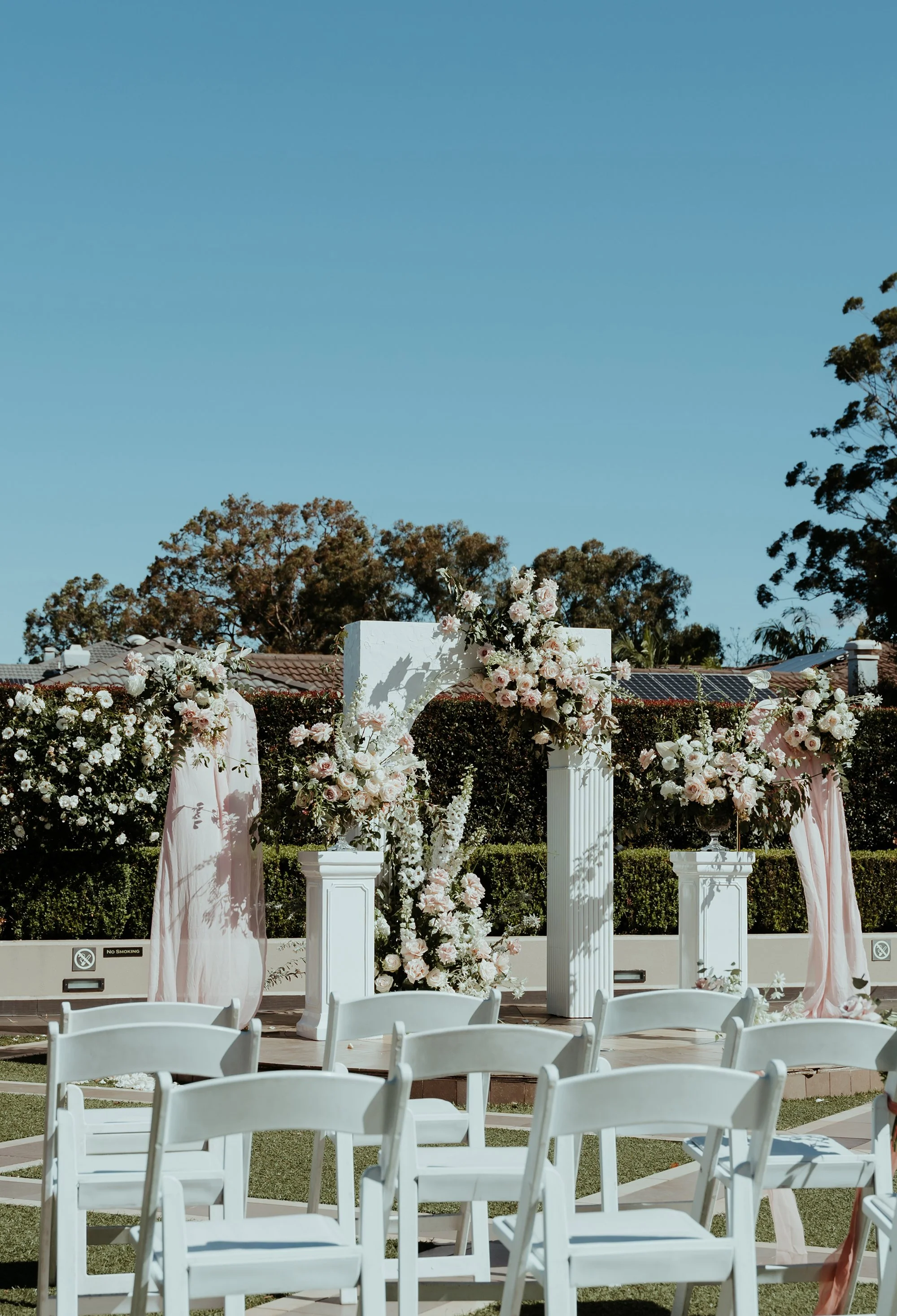 Outdoor wedding setup with white chairs and floral decorations on columns, under a clear blue sky.