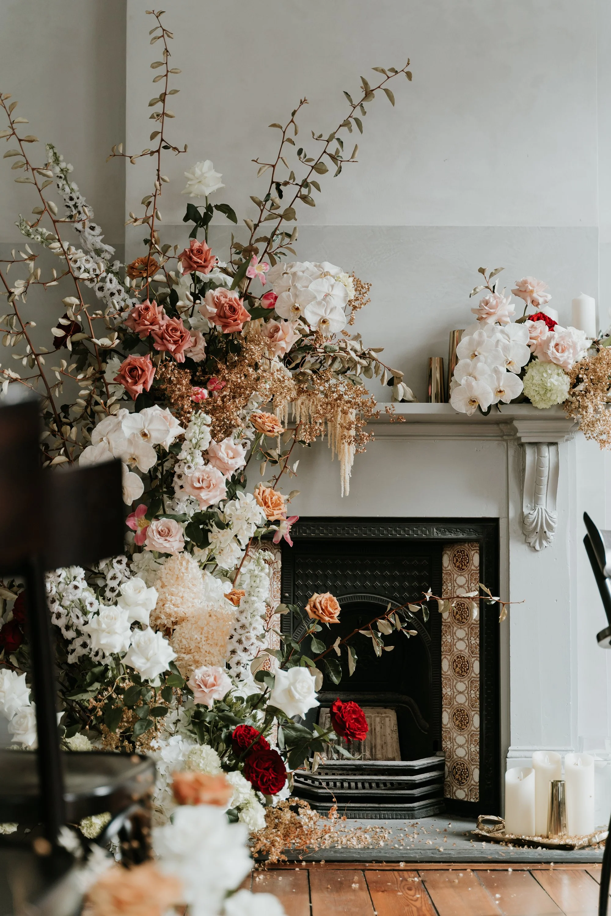 Decorative fireplace with an arrangement of roses and assorted flowers, surrounded by candles on a wooden floor.