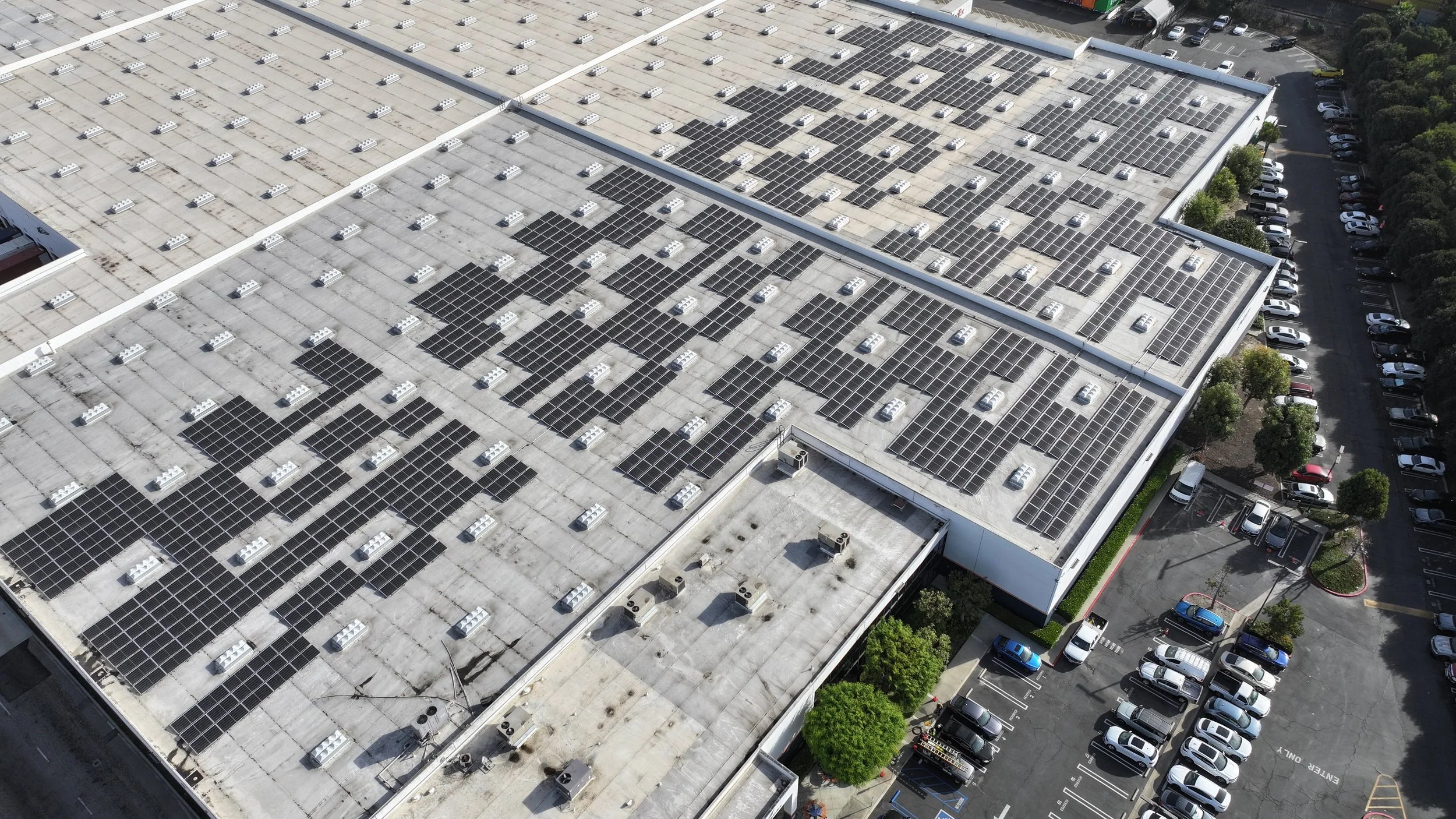 Aerial wide shot of a large commercial warehouse flat roof in Carson, CA covered with hundreds of flat-mounted solar panels showing dust and soiling buildup across multiple array sections