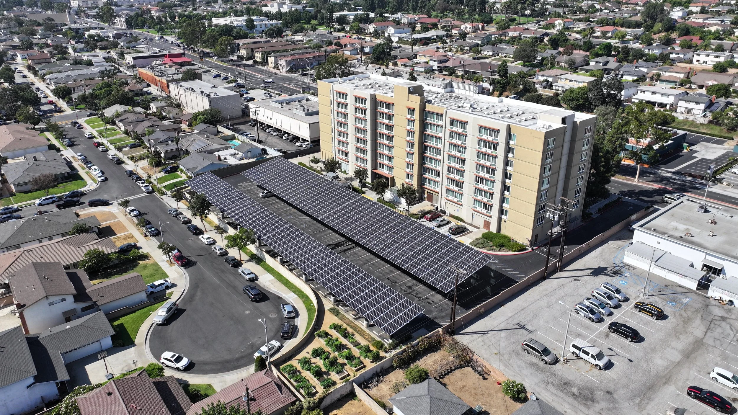 Wide aerial view of a large commercial solar carport array covering a parking lot next to a multi-story building in Montebello, CA, with panels appearing clean and uniform after professional solar panel cleaning service.