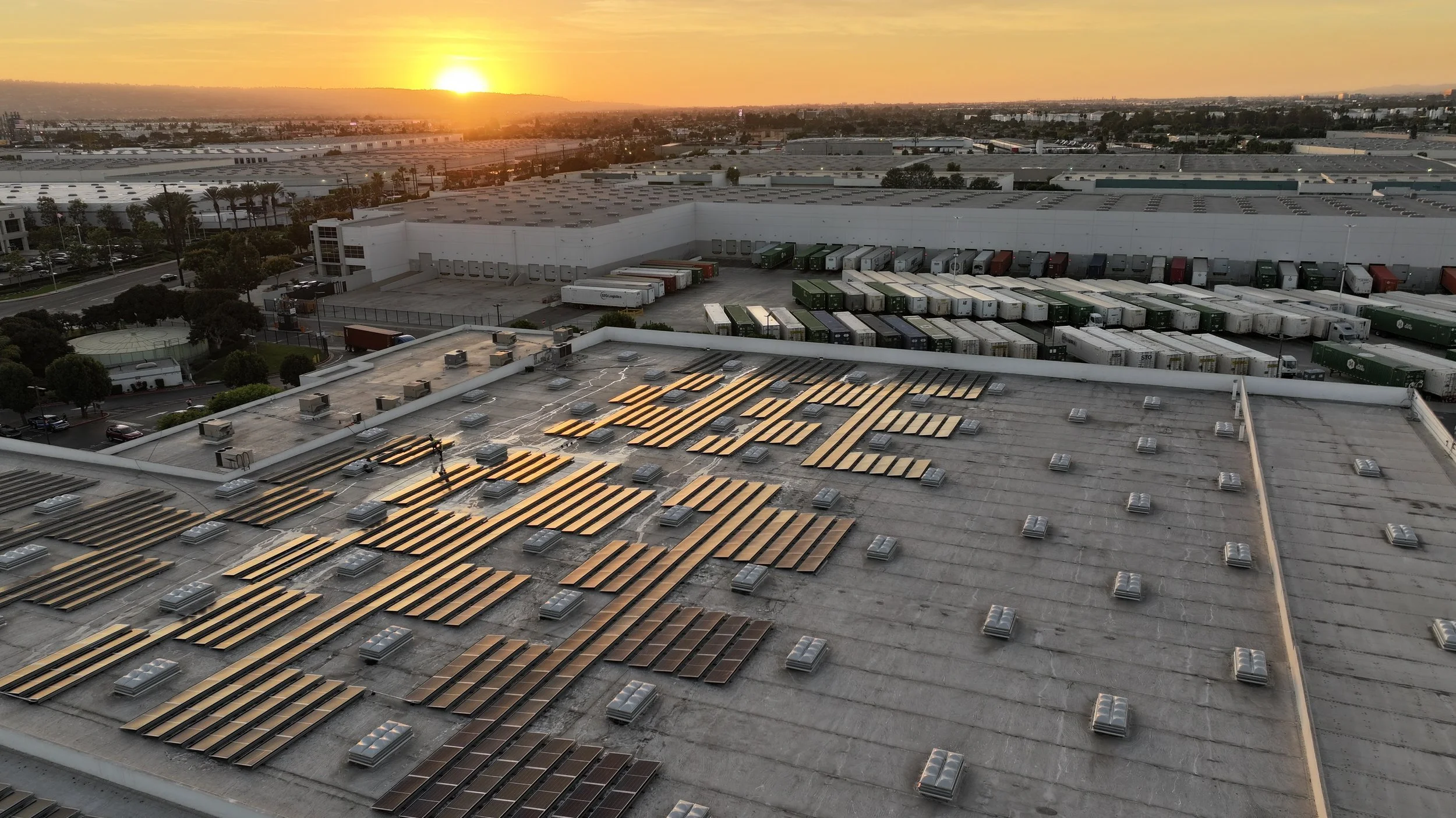 Aerial wide shot at sunset of a large commercial warehouse rooftop in Carson, CA with multiple rows of flat-mounted solar panels showing visible soiling and dust buildup across the array, shot during a Guru Solar Cleaning service visit