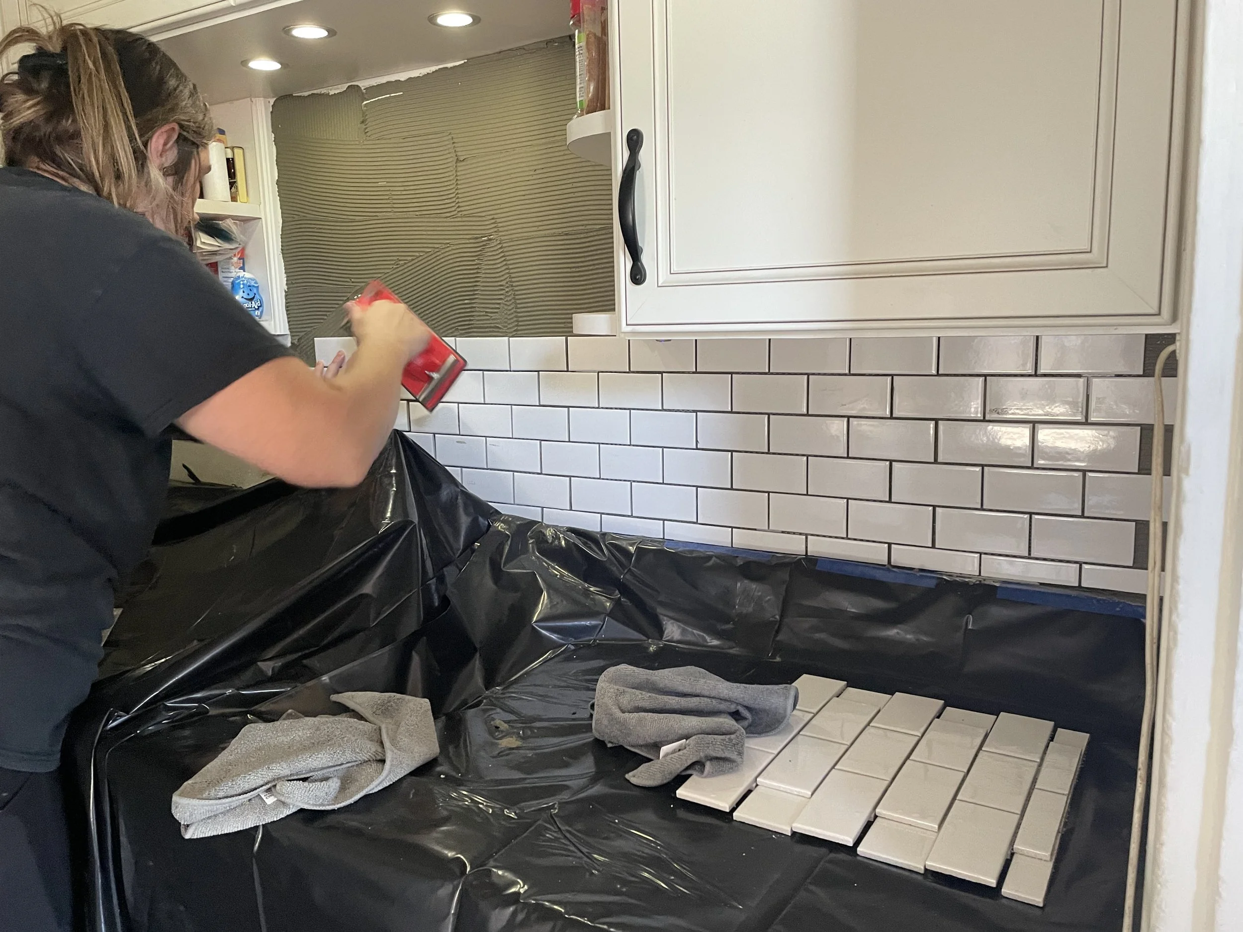 A person working on installing subway tile backsplash in a kitchen, laying out new white tiles over a black plastic-covered surface, with tools and gloves nearby.
