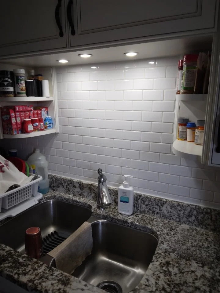 Kitchen sink with a granite countertop, soap dispenser, and a towel hanging on the divider. Overhead lights illuminate the white brick-style backsplash. Open shelves with various containers and boxes are visible on the sides.