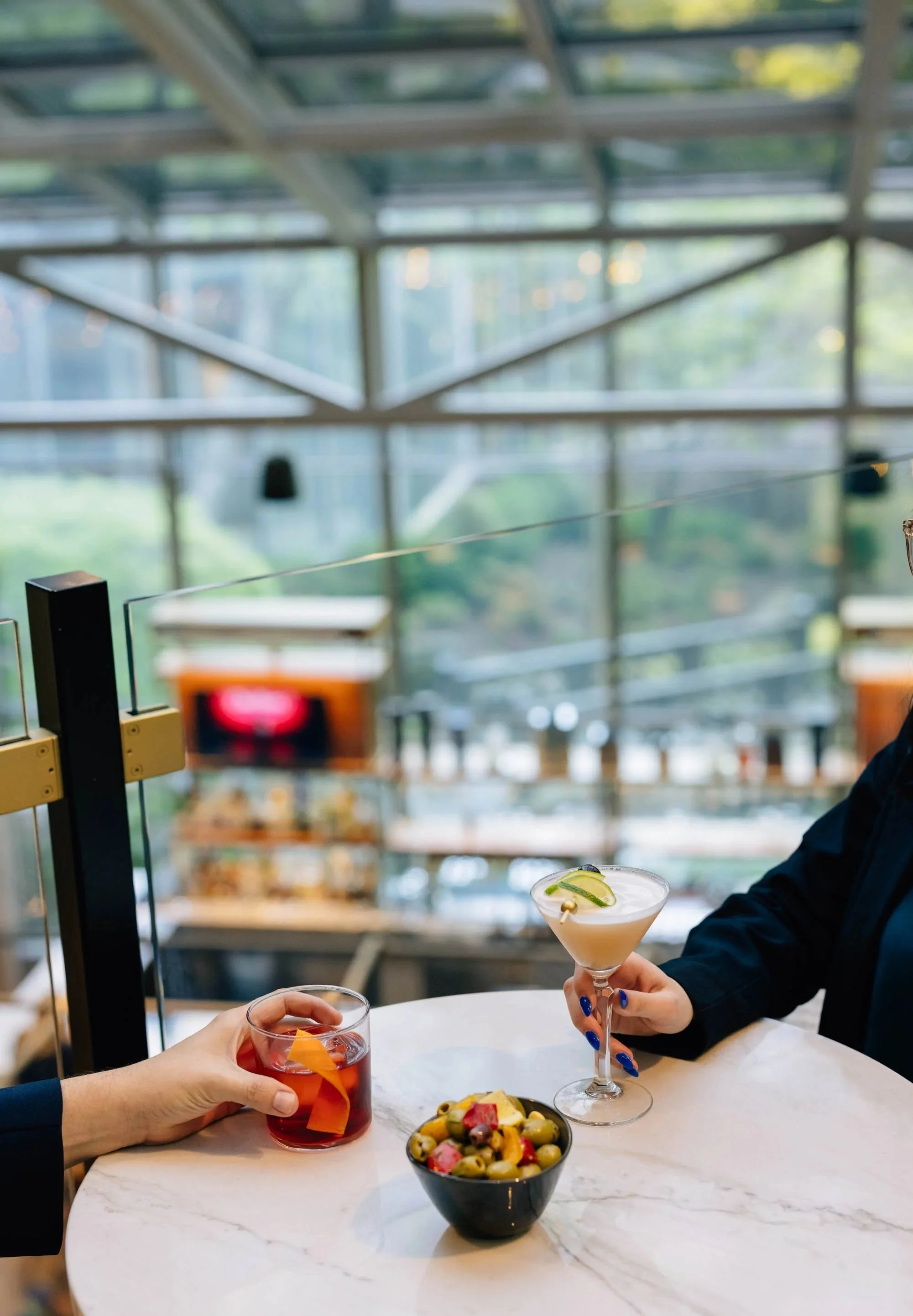 Person holding a cocktail glass with lime garnish, another person holding a red drink with orange peel, a bowl of mixed fruit, two people sit at a marble table inside a glass-walled building.