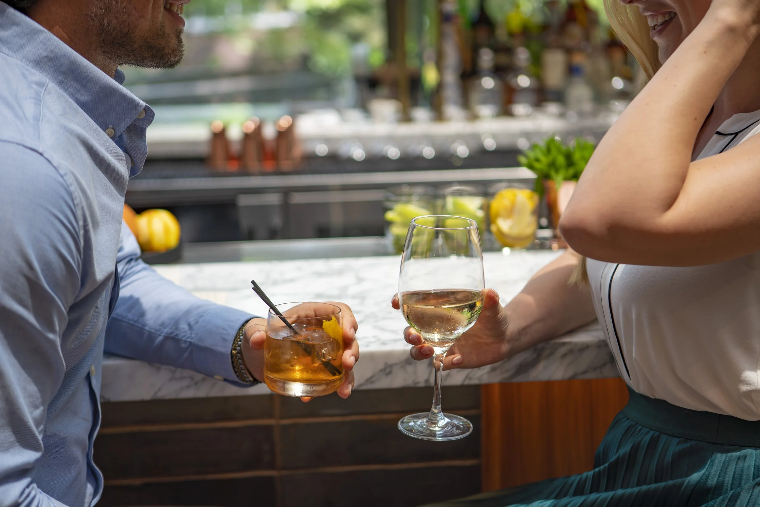 A man and woman enjoying drinks at a bar, with the woman holding a wine glass and the man holding a cocktail glass with a straw and lemon garnish, in a bright, modern setting with greenery and kitchen background.