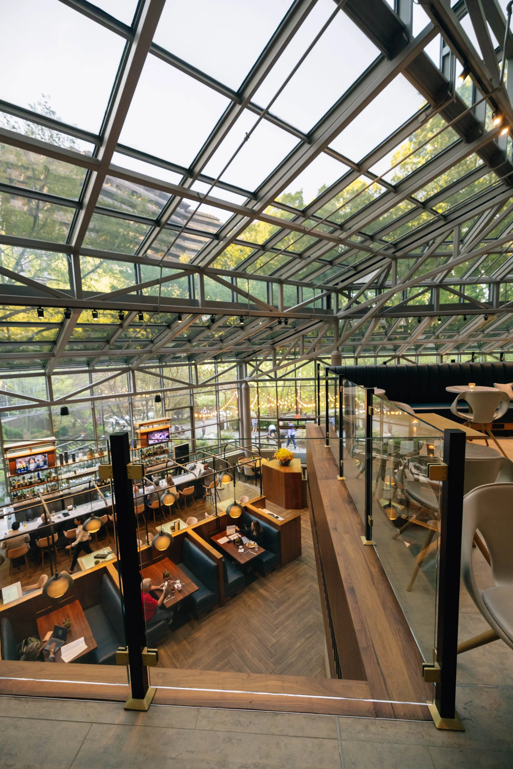 Interior view of a modern restaurant or cafe with a high glass ceiling, showing trees and sky outside. The space has wooden floors, booth seating, tables with chairs, people dining, and decorative hanging lights.