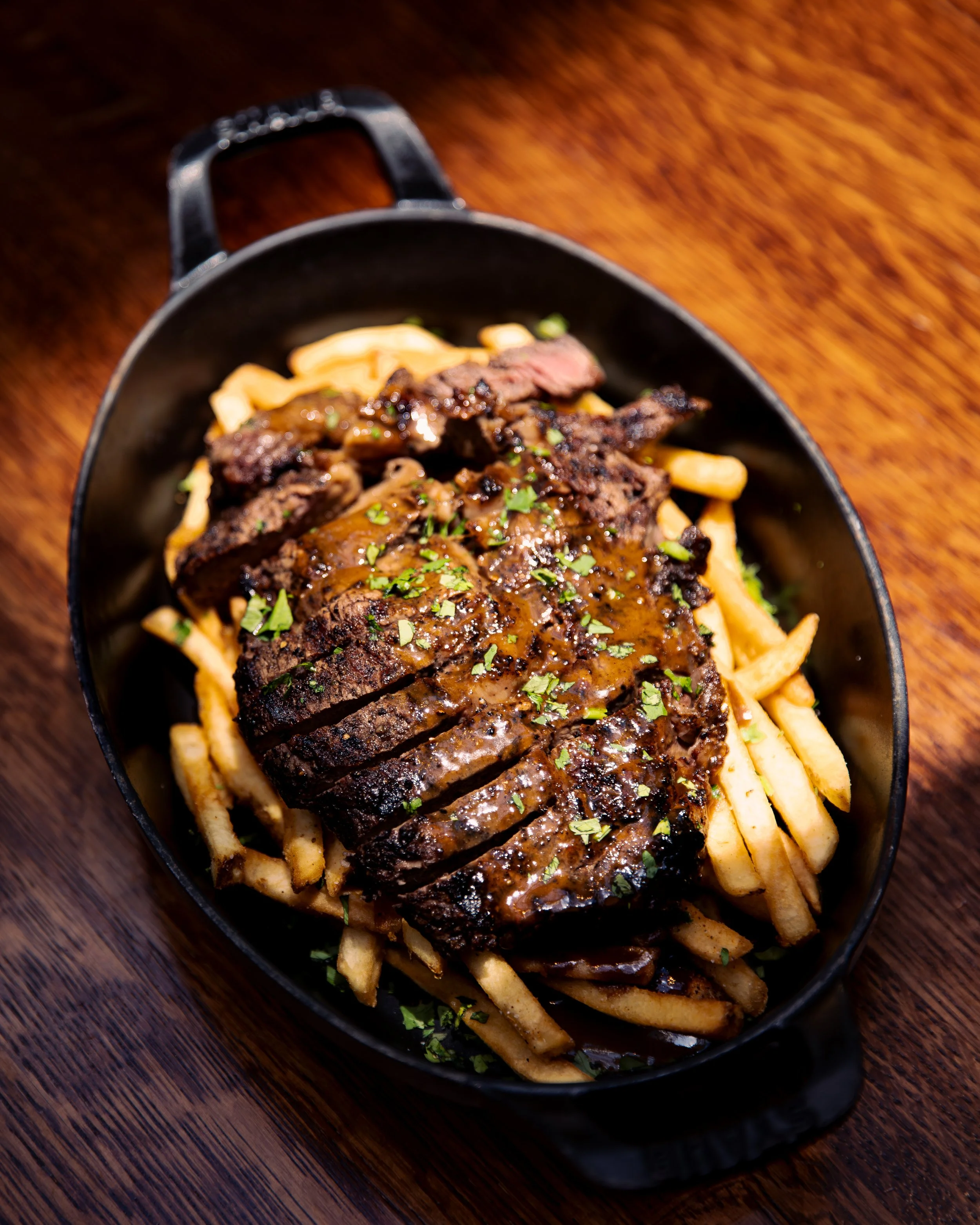 A black oval dish containing crispy French fries topped with slices of grilled steak and beef gravy, garnished with chopped parsley, on a wooden table.