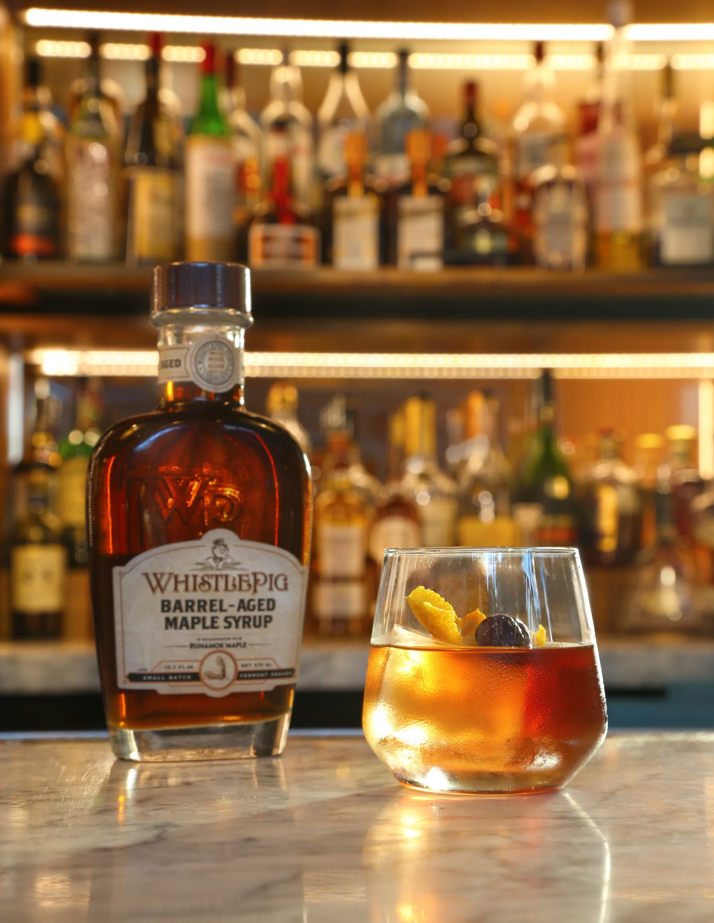 A bottle of WhistlePig Barrel-Aged Maple Syrup and a glass of maple syrup with garnishes on a marble surface, with a blurred bar background.
