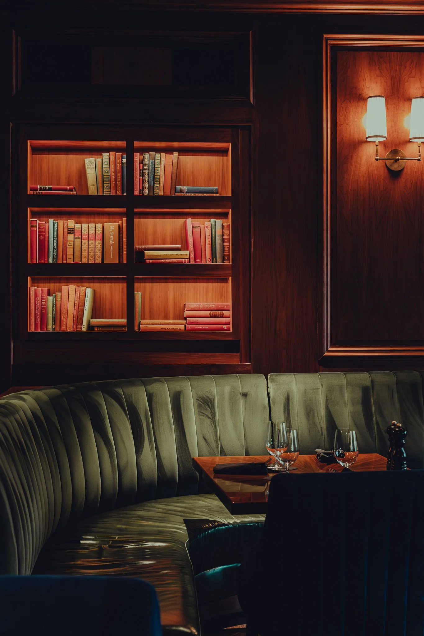 A cozy restaurant corner with dark wood paneling, a built-in bookshelf filled with colorful books, a wall-mounted light fixture, a curved green velvet banquette, and a table set with glasses, knives, and a pepper grinder.
