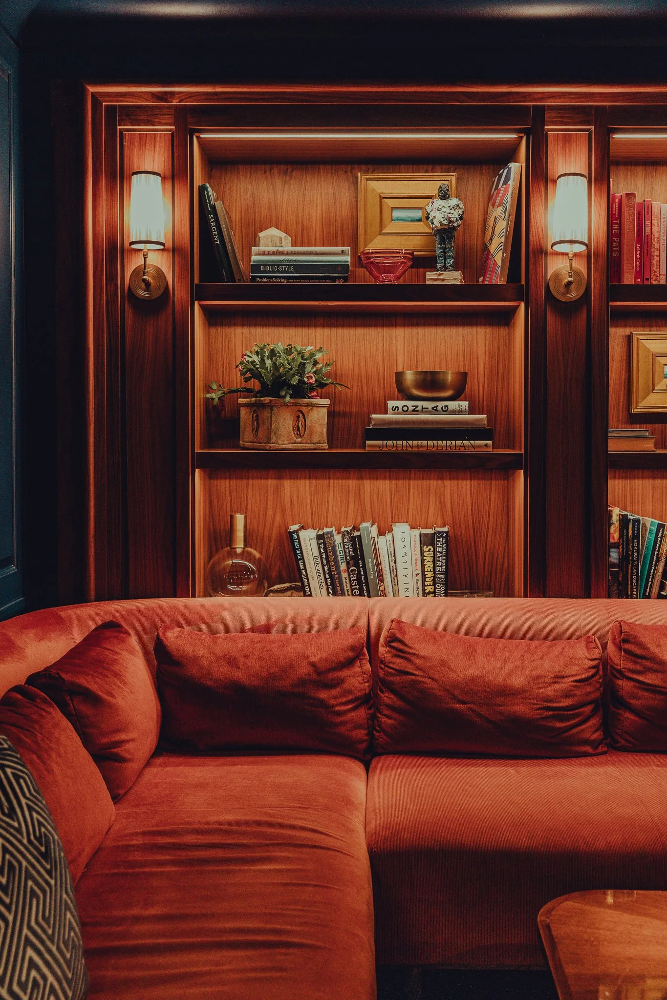 A cozy living room with a red velvet sofa, wooden bookshelf with books and decorative items, and wall-mounted lamps on ornate wooden paneling.