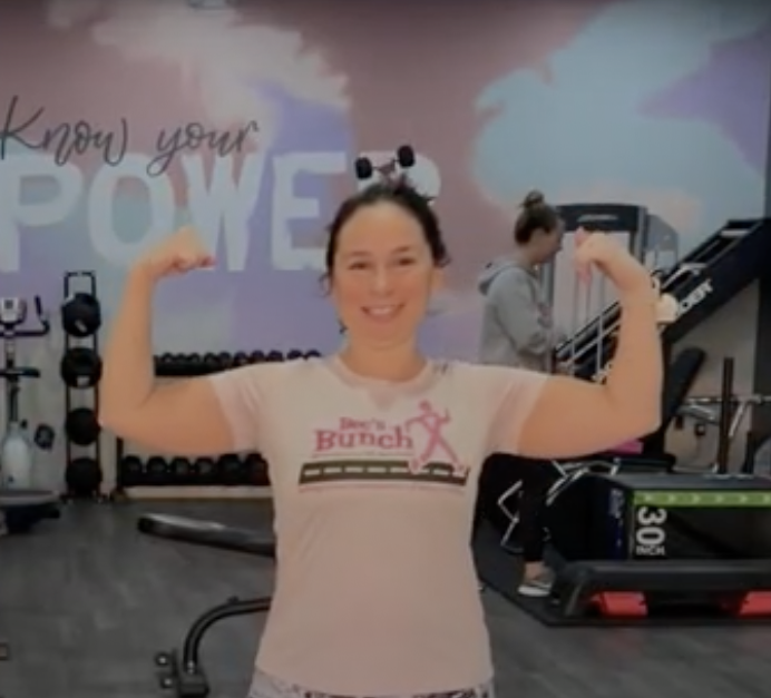 A woman smiling and flexing her biceps in a gym, with a background that includes exercise equipment and the words 'Know your Power' on the wall.