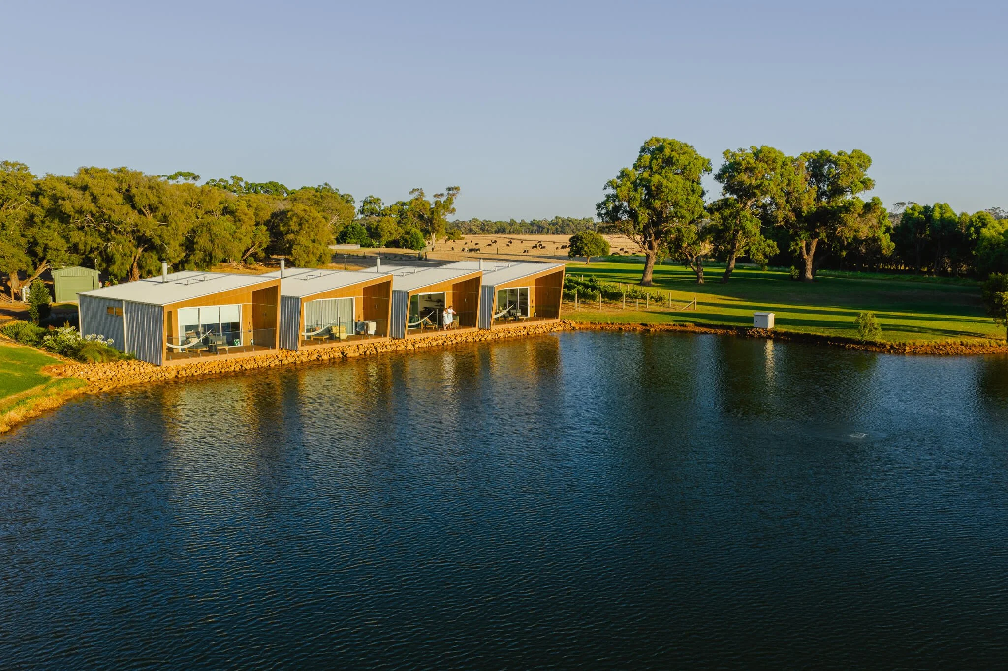 Modern boathouse with large glass windows and sloped roof, situated along the edge of a calm lake, surrounded by lush green trees and open grassy areas in the background, under a clear blue sky near Margaret River.