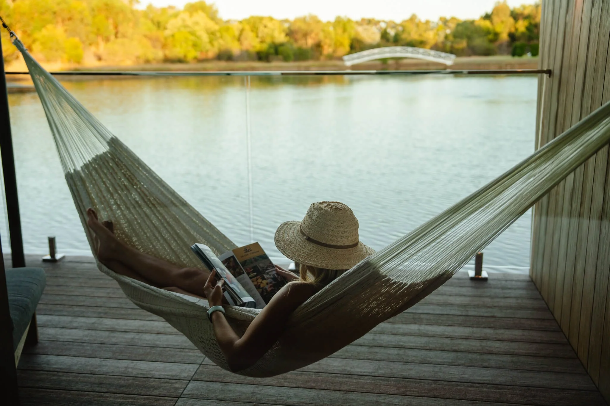 Person relaxing in a hammock on a porch by a lake, reading a magazine, with trees and a bridge in the background.