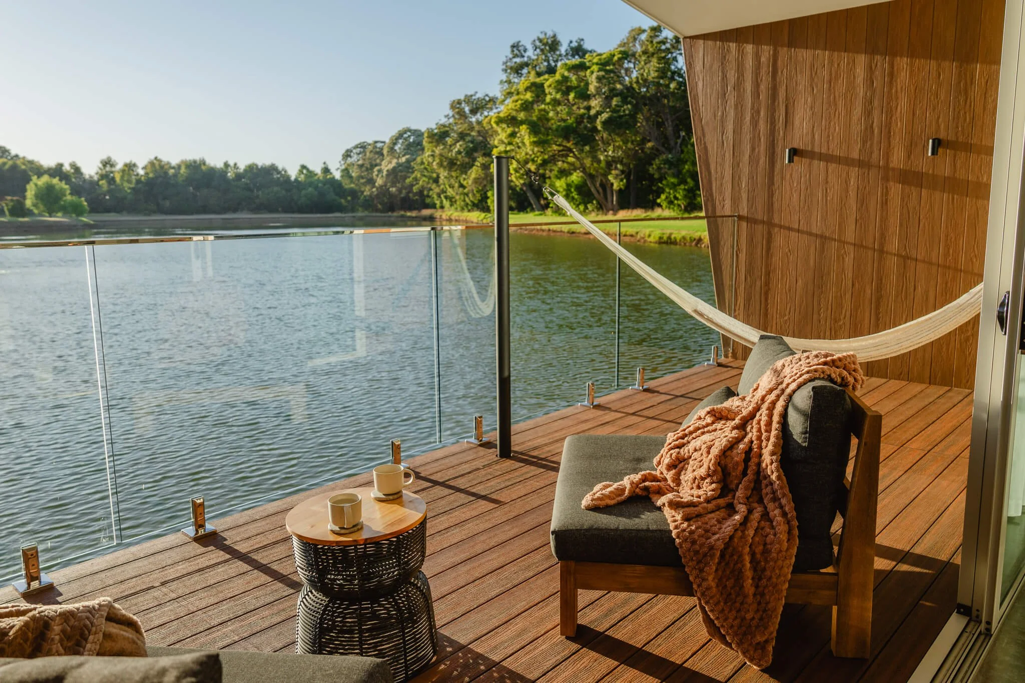A balcony overlooking a body of water with trees in the background. The balcony has glass railings and wooden flooring, with a hammock, a sofa with a blanket, and a small table with two cups.