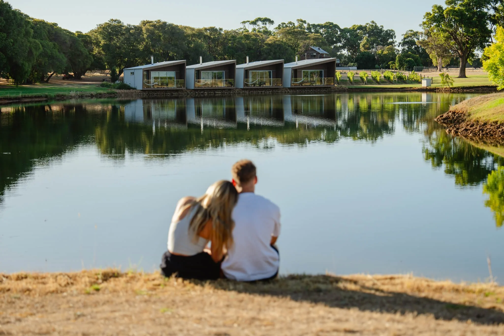 A couple sitting by a pond, gazing at modern houses across the water, with green trees and a sunny sky in the background.