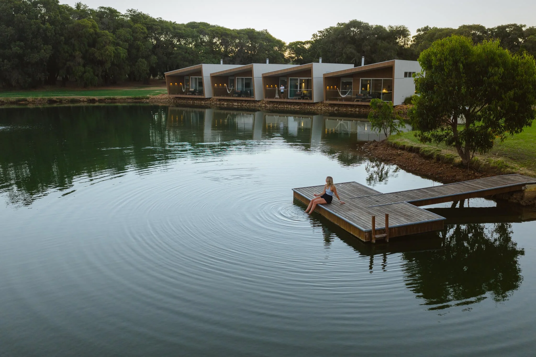 A woman sitting on a wooden dock by a lake, with modern cabins in the background and trees surrounding the area during the evening.