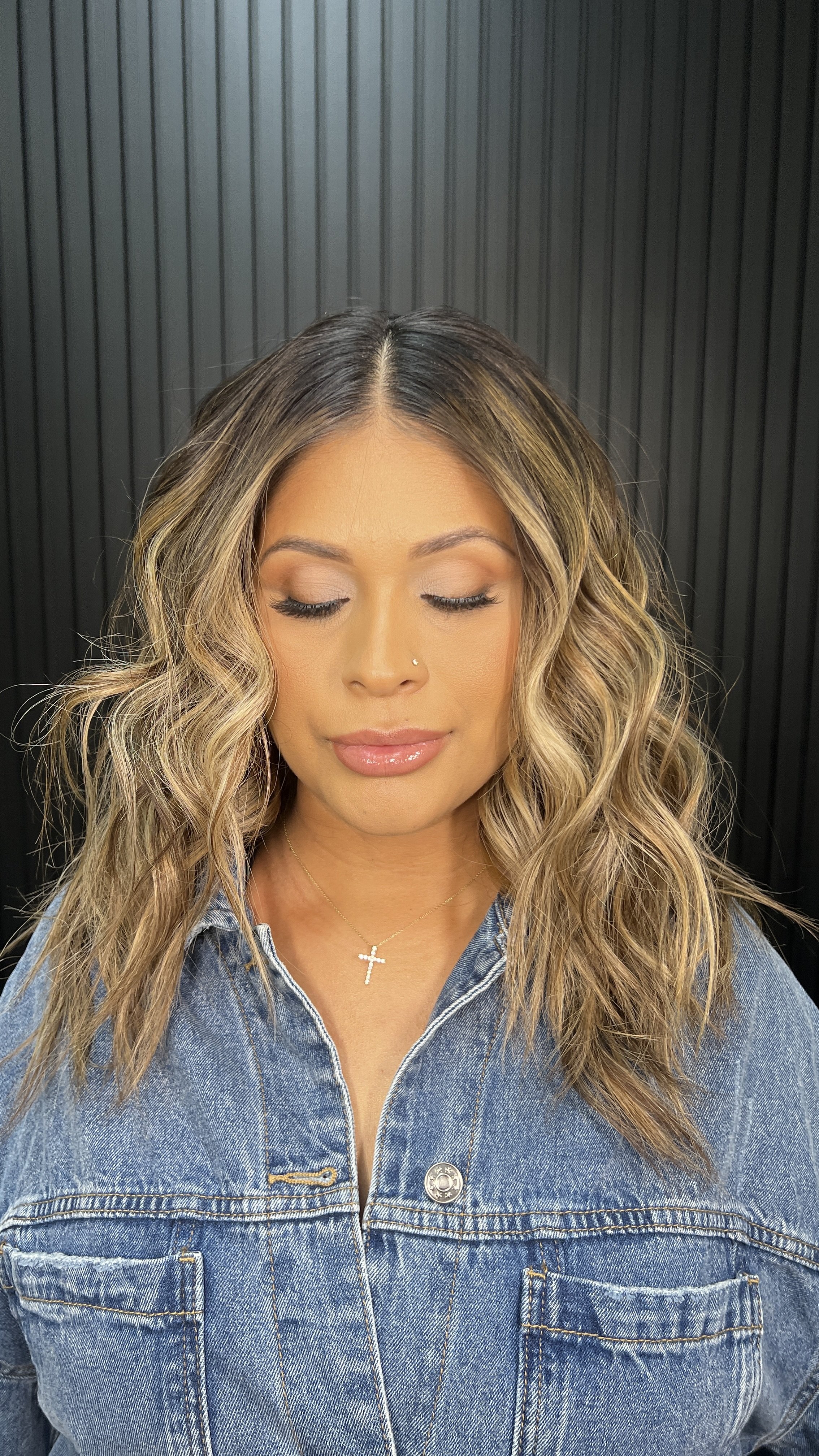 A woman with curly, shoulder-length hair wearing a denim jacket and a cross necklace, with her eyes closed and a neutral expression, standing in front of a black, ribbed background.