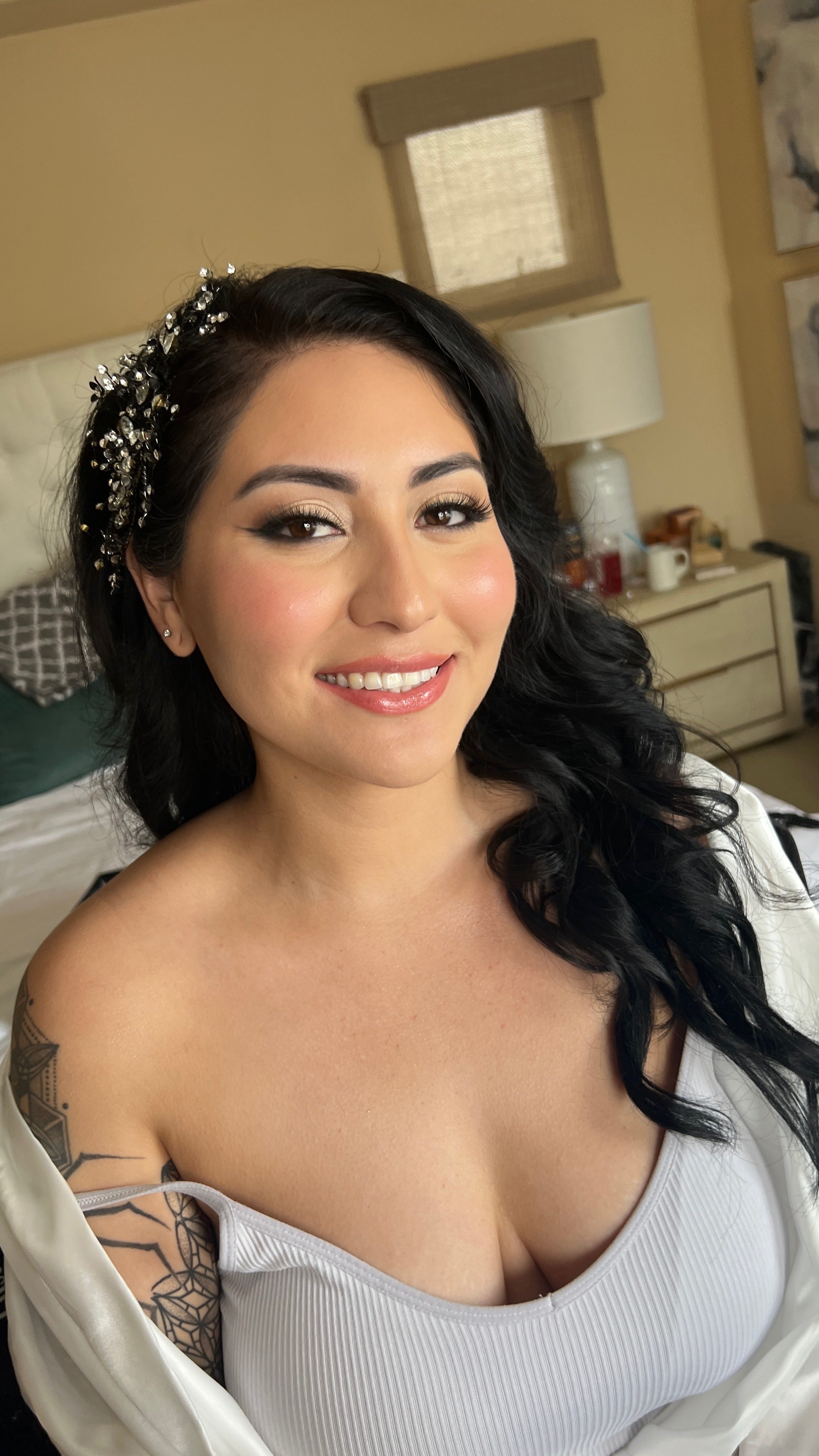 A smiling woman with long black curly hair, wearing a white off-shoulder top and a decorative headband with crystals, posing in a bedroom.