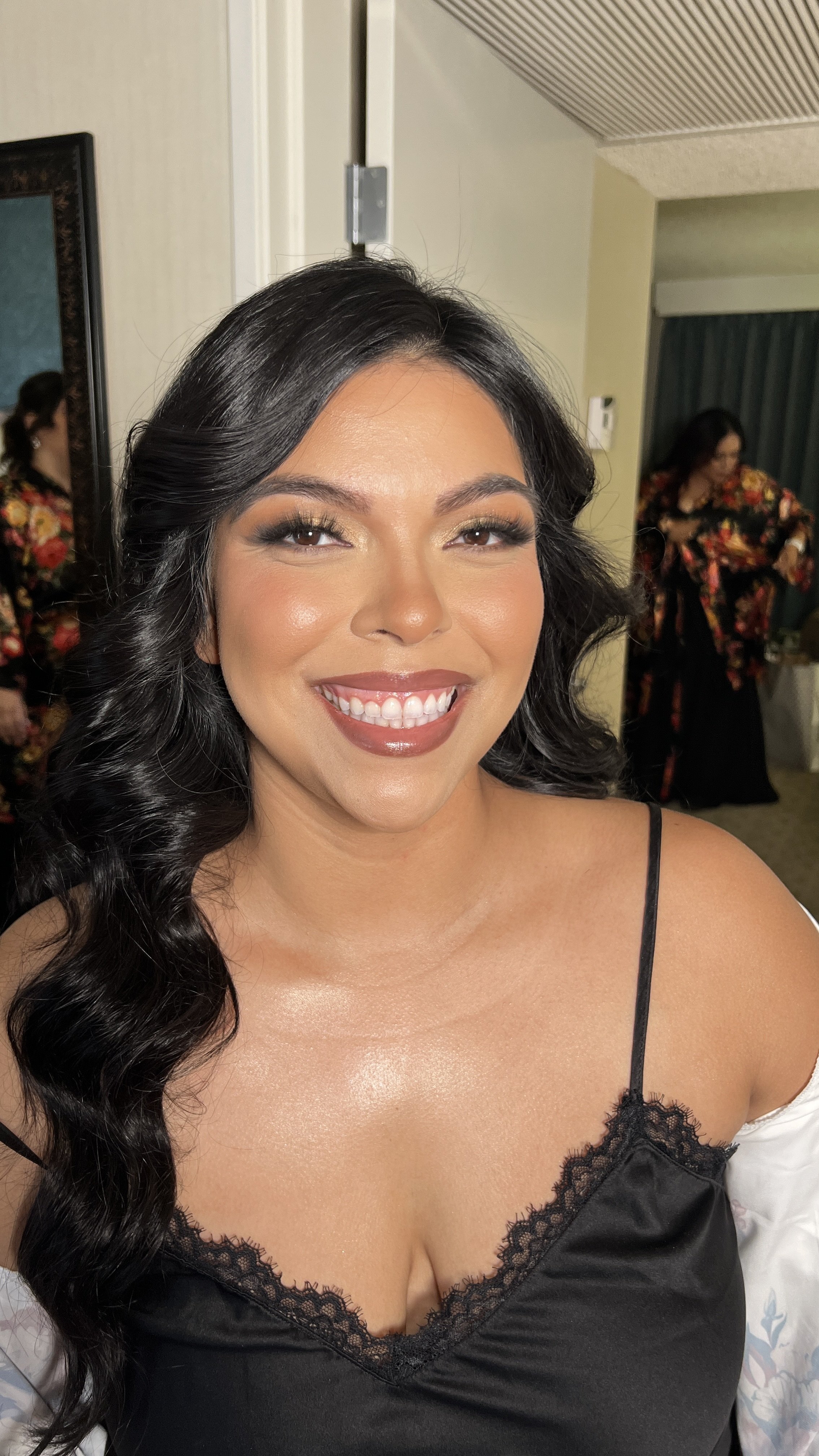 A smiling woman with long black hair, wearing makeup and a black spaghetti strap top, standing indoors with other women in the background.