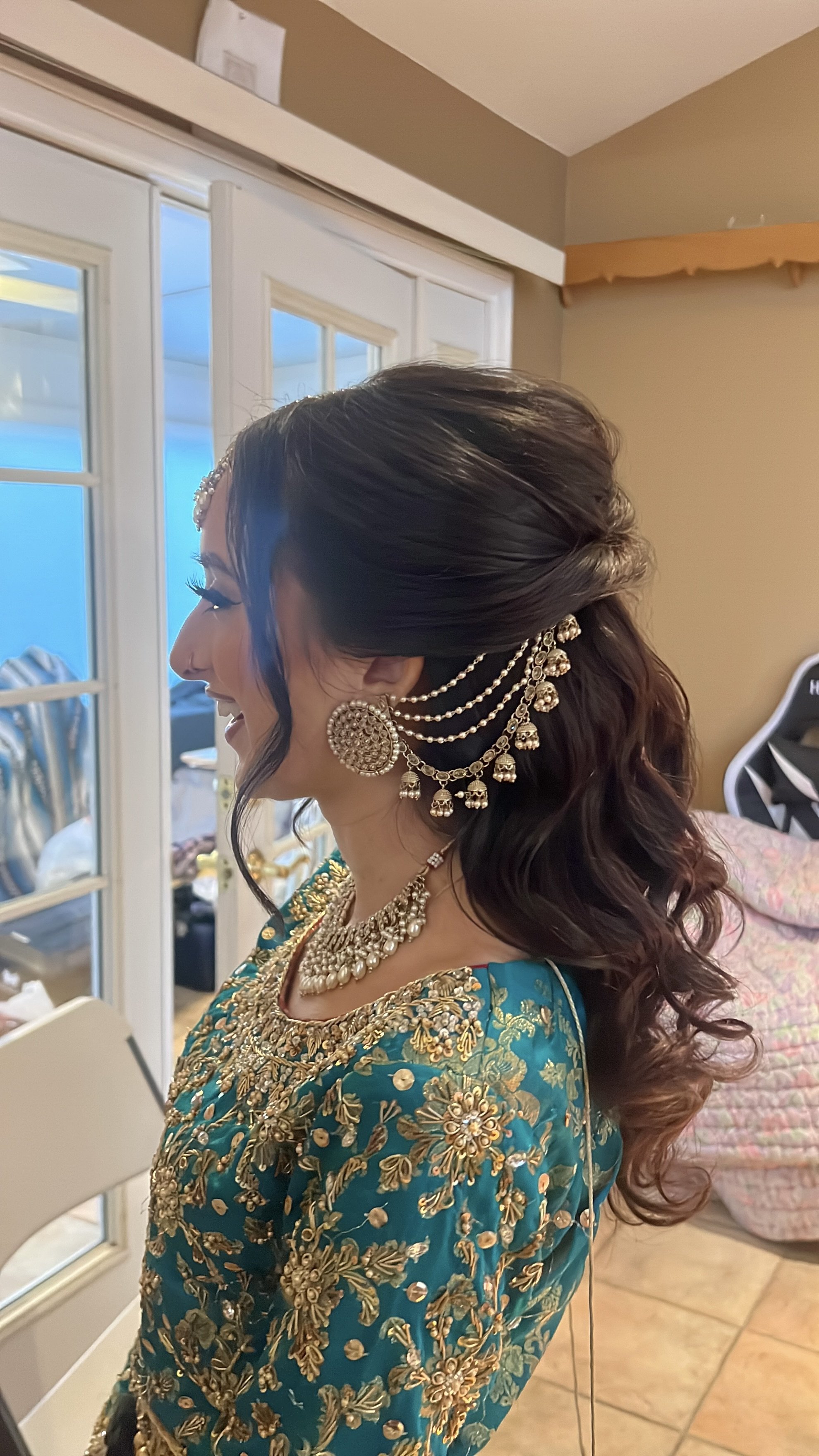 A woman in traditional Indian attire with ornate gold embroidery, wearing elaborate jewelry including earrings, necklaces, and a hair accessory, standing indoors with her eyes closed and smiling.