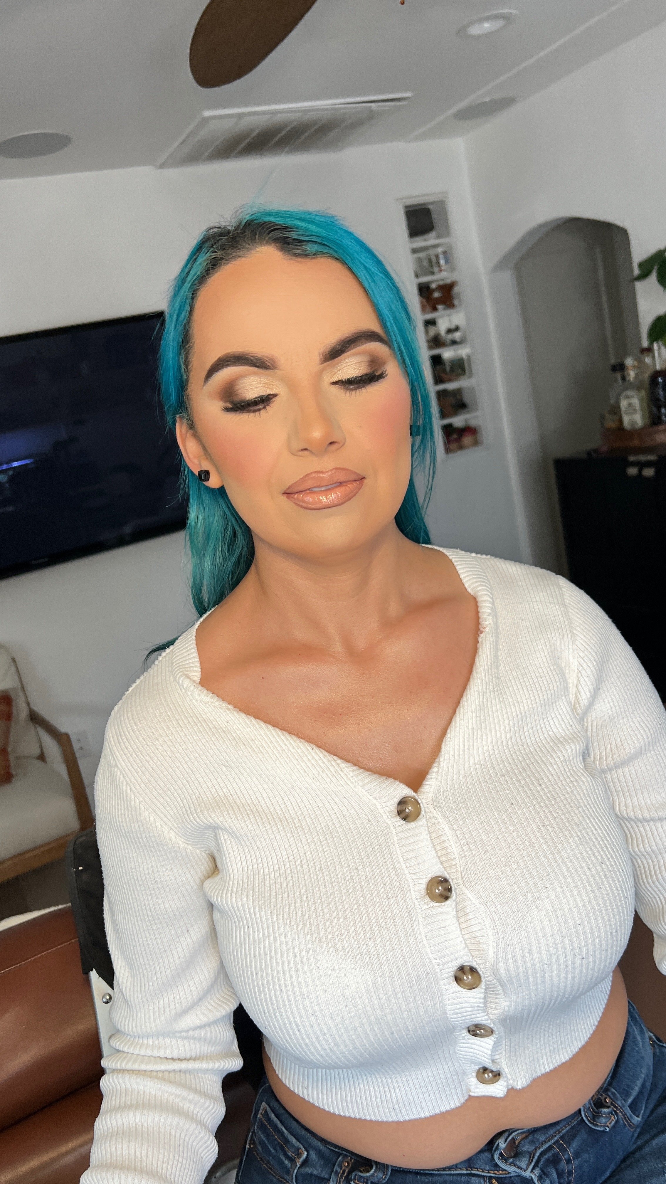 A woman with bright blue hair and makeup, wearing a white ribbed button-up sweater and blue jeans, standing indoors with eyes closed.