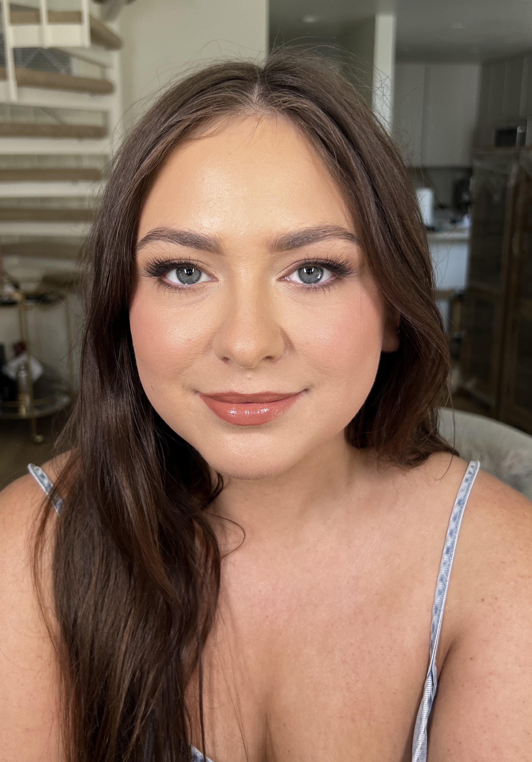 Close-up of a young woman with long, wavy brown hair, light complexion, blue eyes, wearing makeup, and a thin-strapped top, smiling softly in an indoor setting.