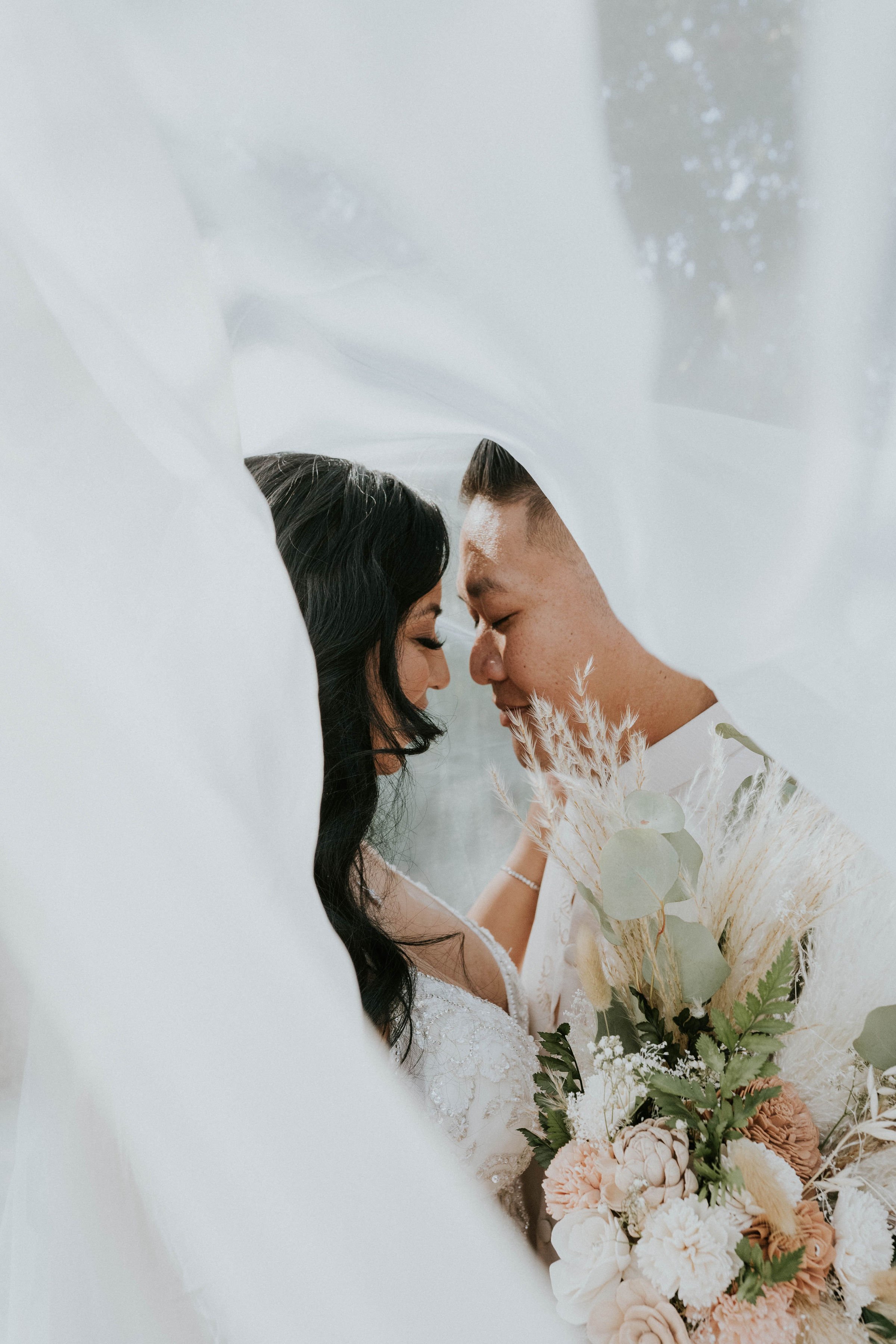 A couple on their wedding day captures an intimate moment through a white fabric or veil, with the bride holding a bouquet of flowers and both with their eyes closed touching foreheads.