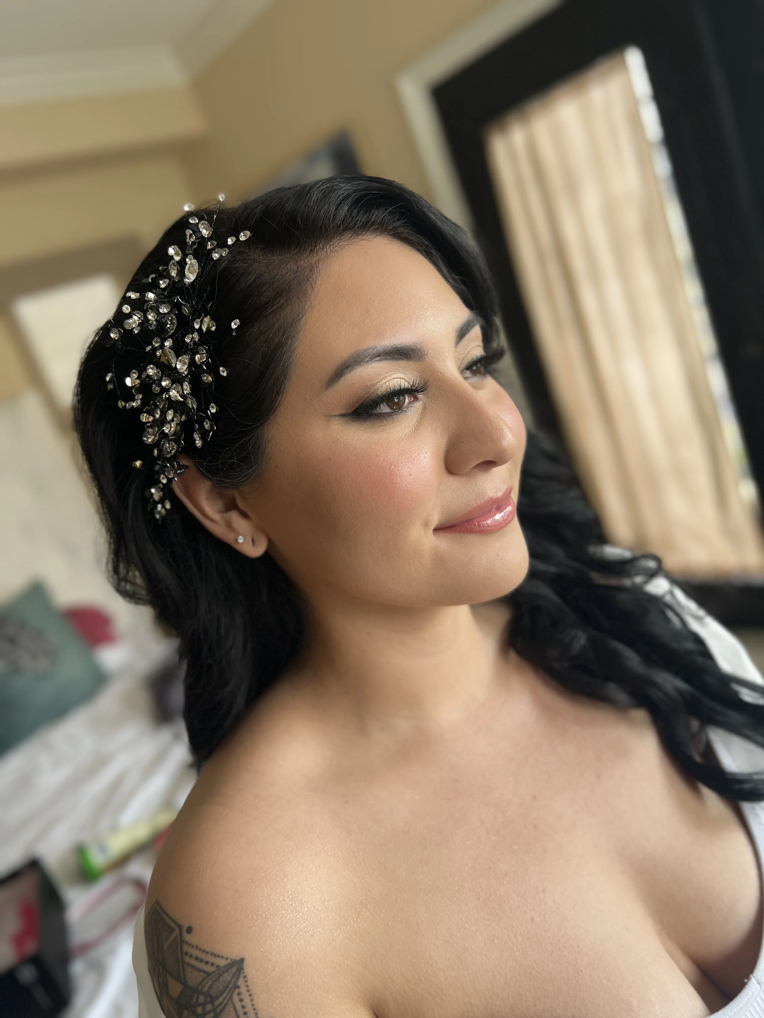 A woman with black hair styled in waves, wearing a decorative hairpiece with rhinestones, is looking off to the side with makeup and a tattoo on her shoulder visible. newport beach bridal makeup and hair