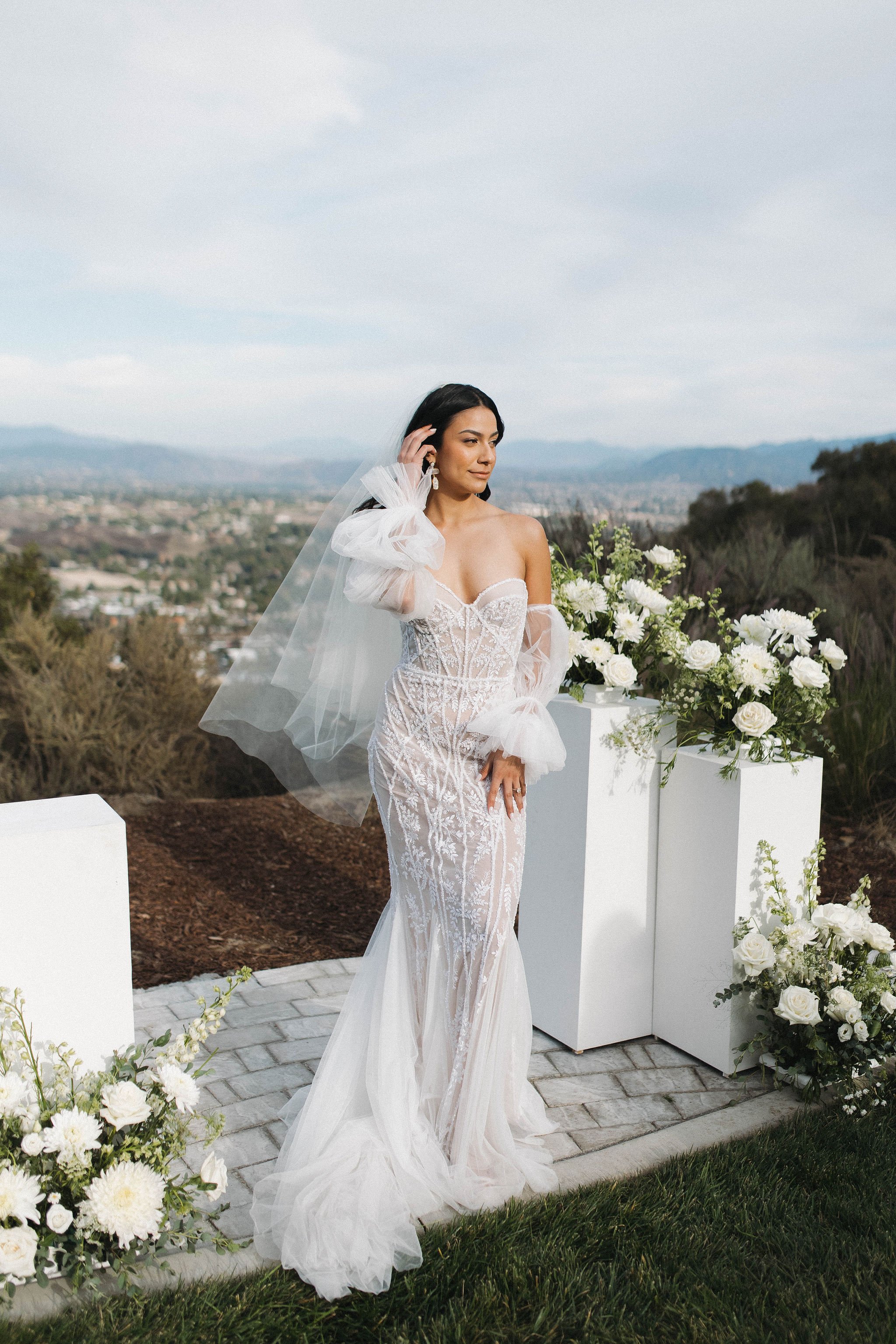 A bride in a detailed white wedding dress with tulle sleeves standing outdoors near flower arrangements with a scenic landscape in the background.