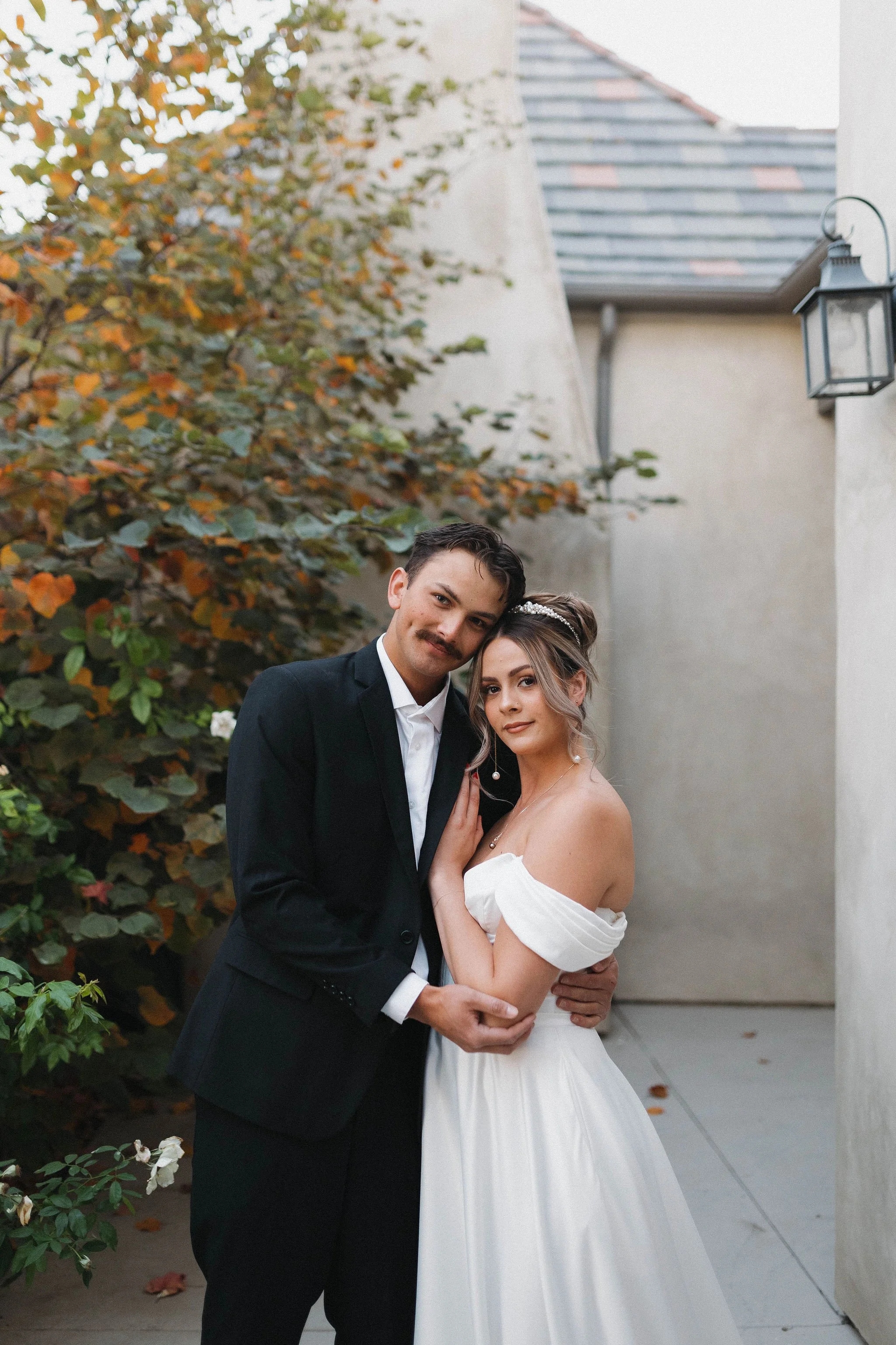 A couple in wedding attire standing close together outdoors, with trees and a building in the background. romantic bridal glam in temecula, california
