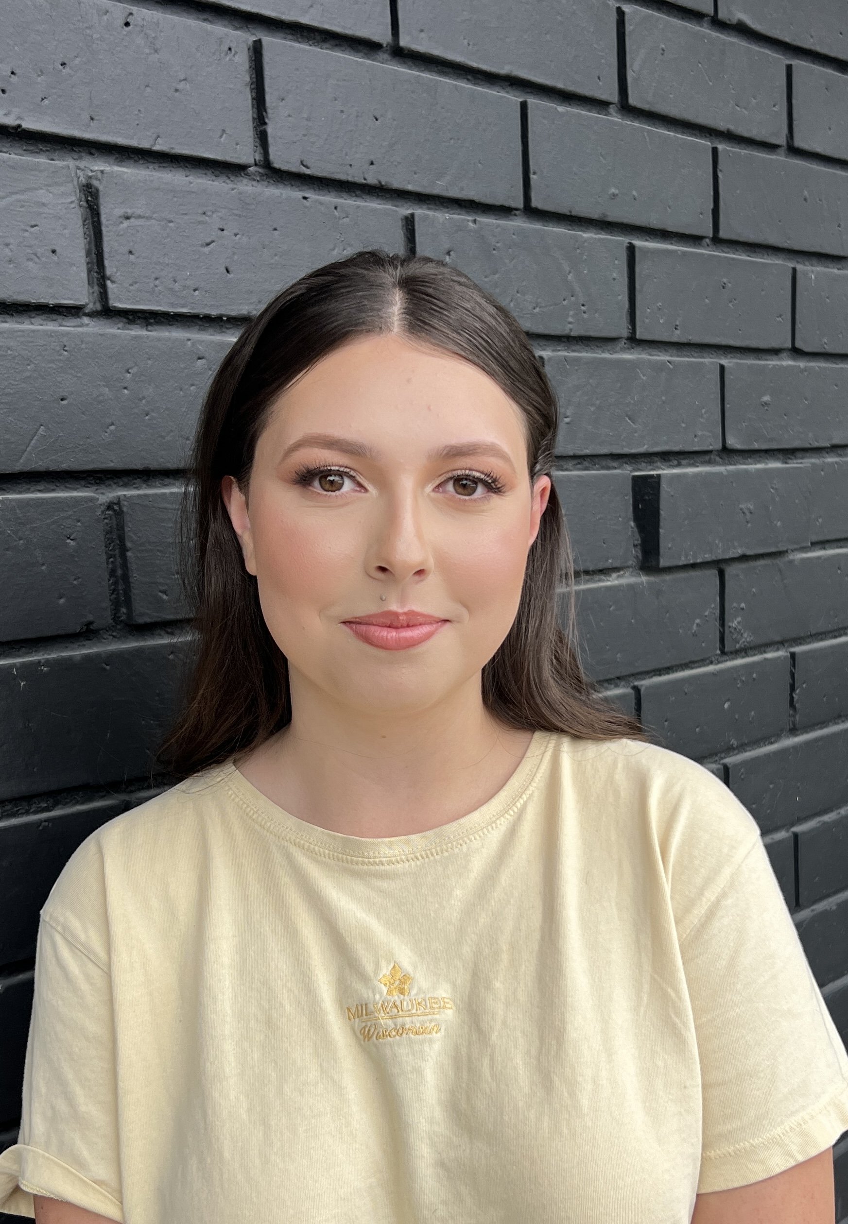 A young woman with long brown hair and light makeup, wearing a yellow T-shirt, standing against a black brick wall.