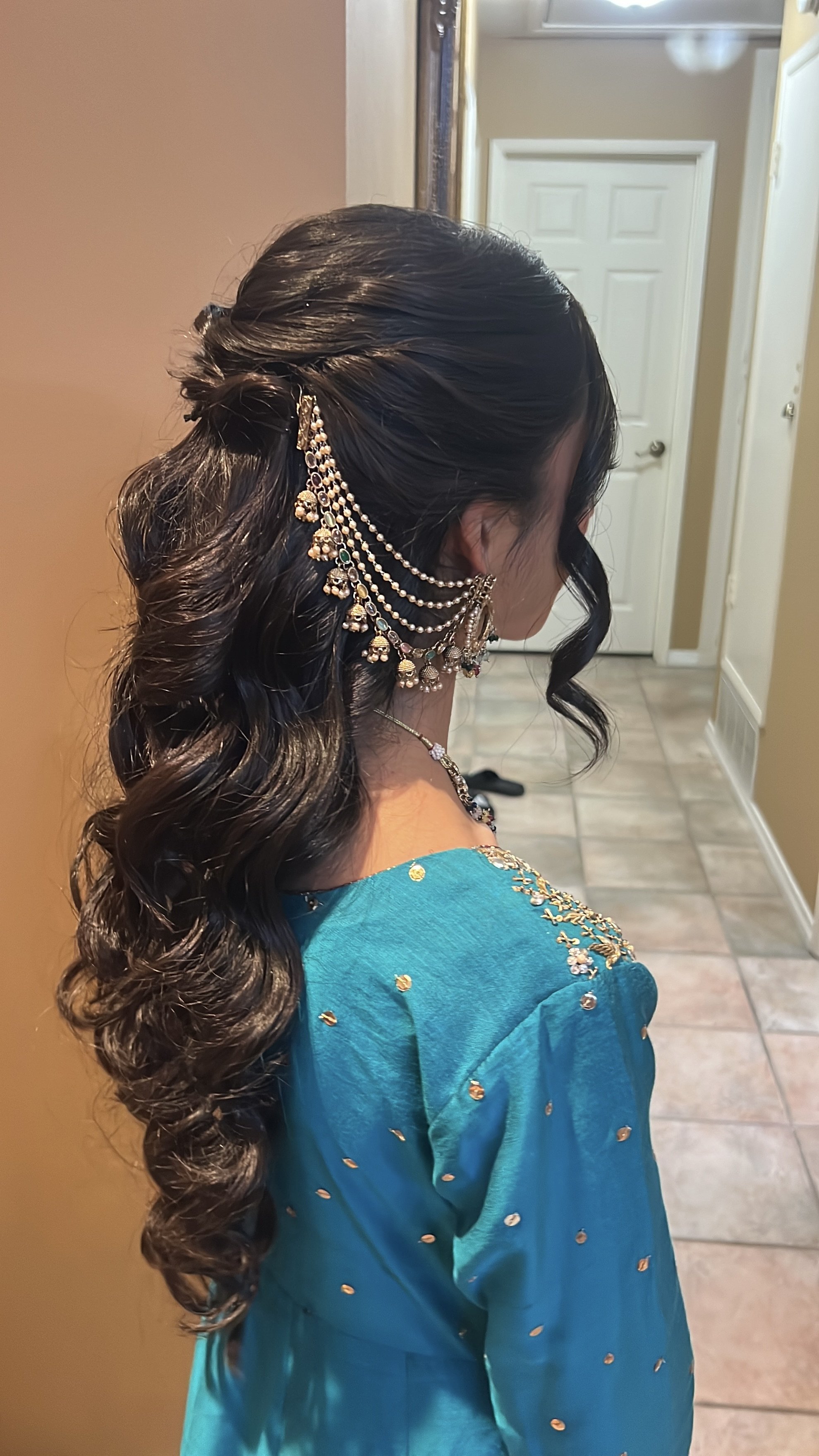 A woman with dark, wavy hair styled in loose curls, wearing traditional Indian jewelry and a turquoise dress with gold embellishments, standing in a doorway.