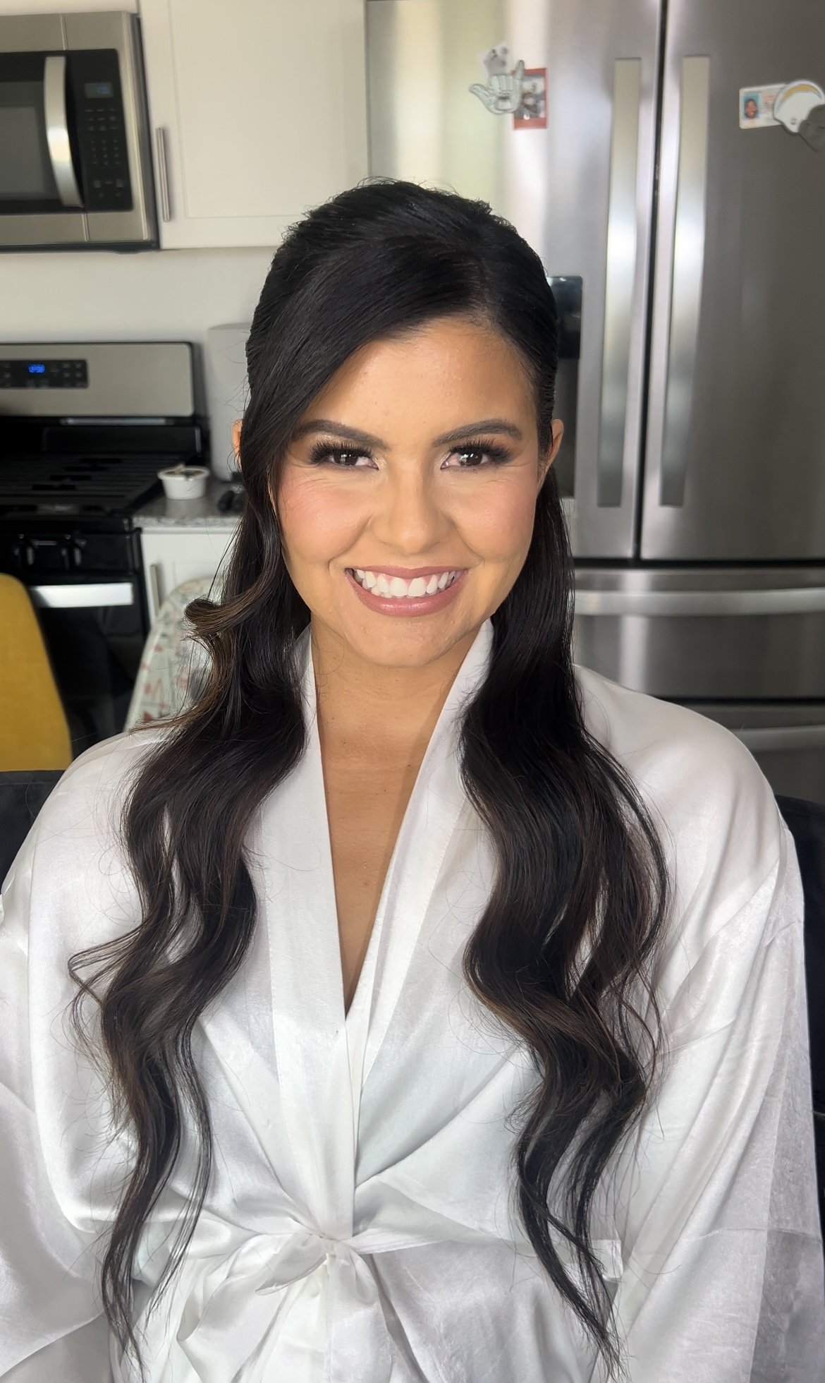 A woman with long, dark wavy hair smiling in a kitchen, wearing a white robe.
