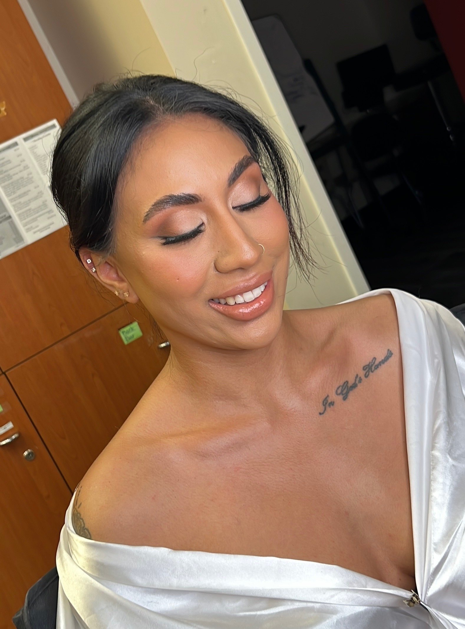 A woman with dark hair and makeup smiling with closed eyes, wearing a white satin gown and displaying a tattoo on her chest that reads "In God's Hand."