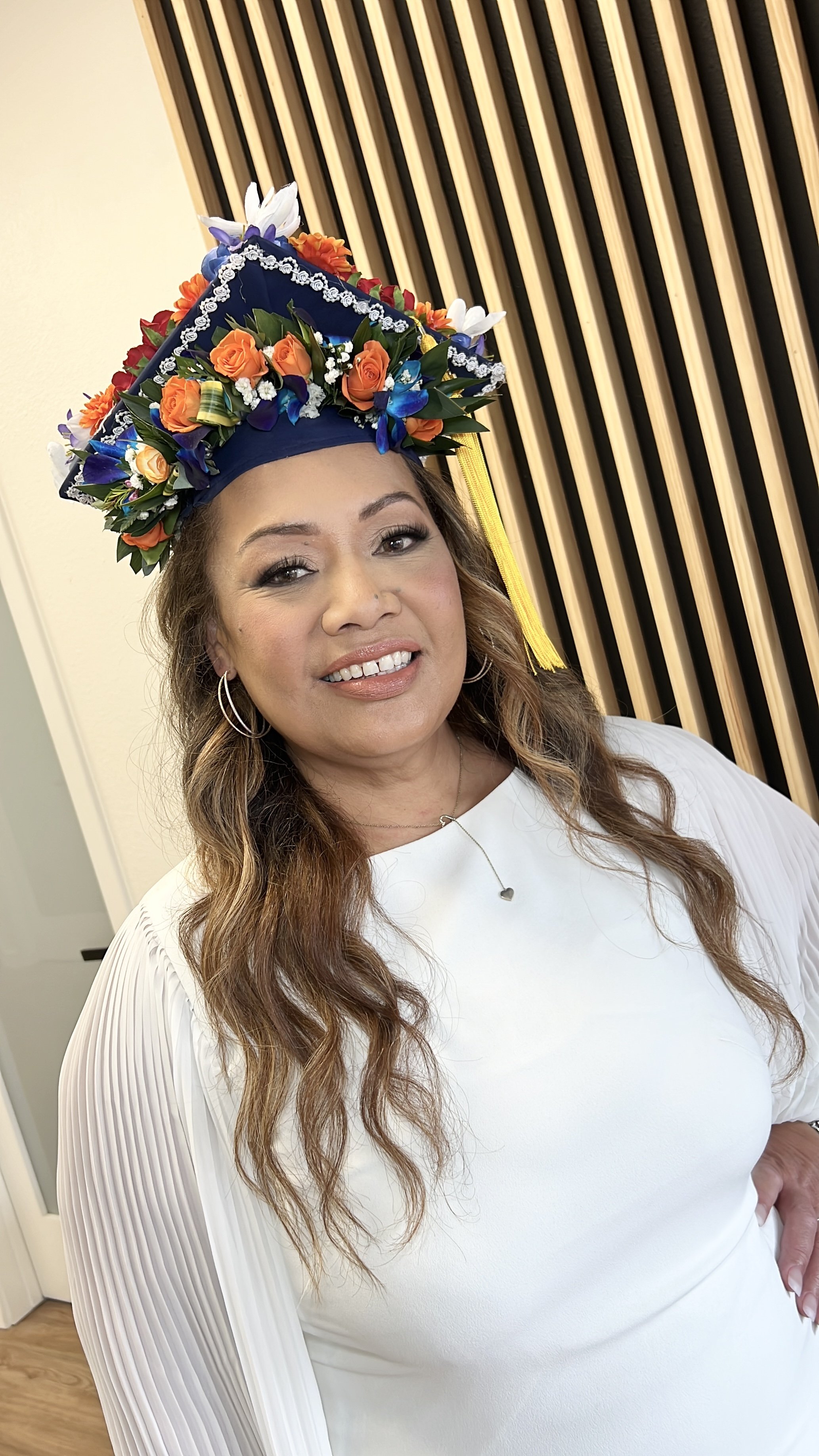 A woman smiling wearing a white dress and a colorful floral cap with orange, purple, white flowers, and a yellow tassel.