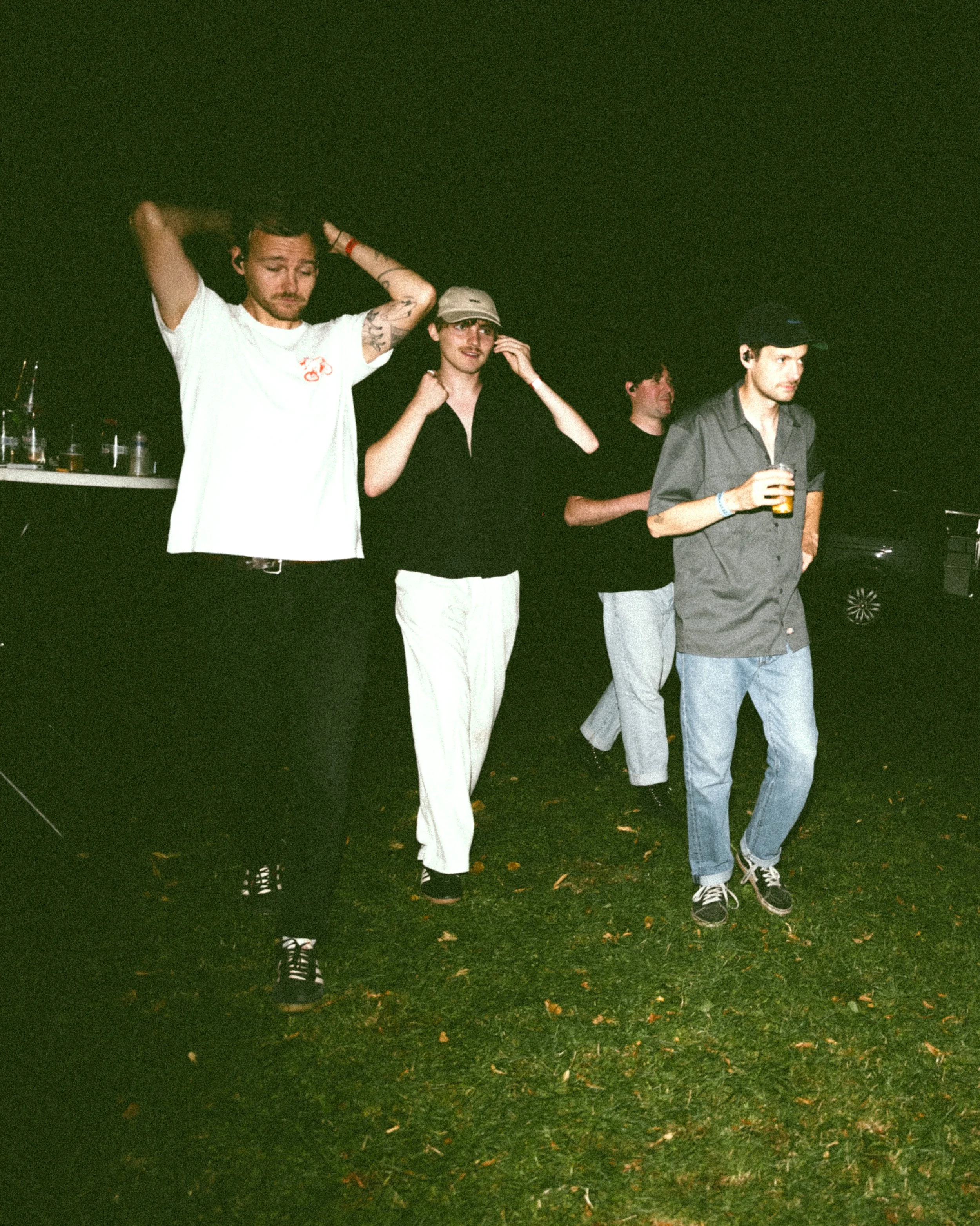 Four men standing on a grassy area at night, with one holding a drink. The first man from the left has tattoos and is adjusting his hair. The second man is wearing a light-colored hat and black shirt, touching his face. The third man is dressed in bl