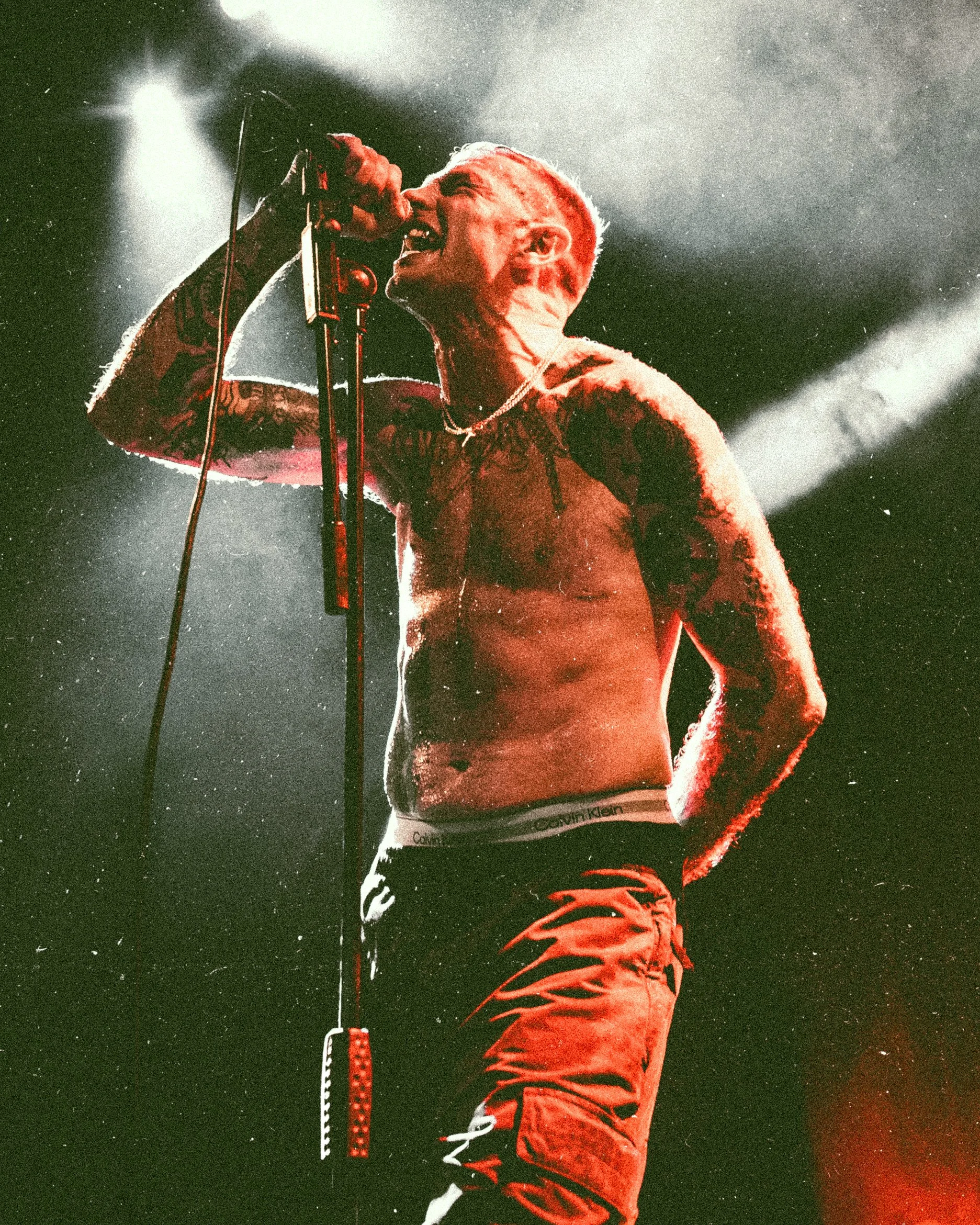 A shirtless male singer with tattoos singing into a microphone on stage, under dramatic lighting.
