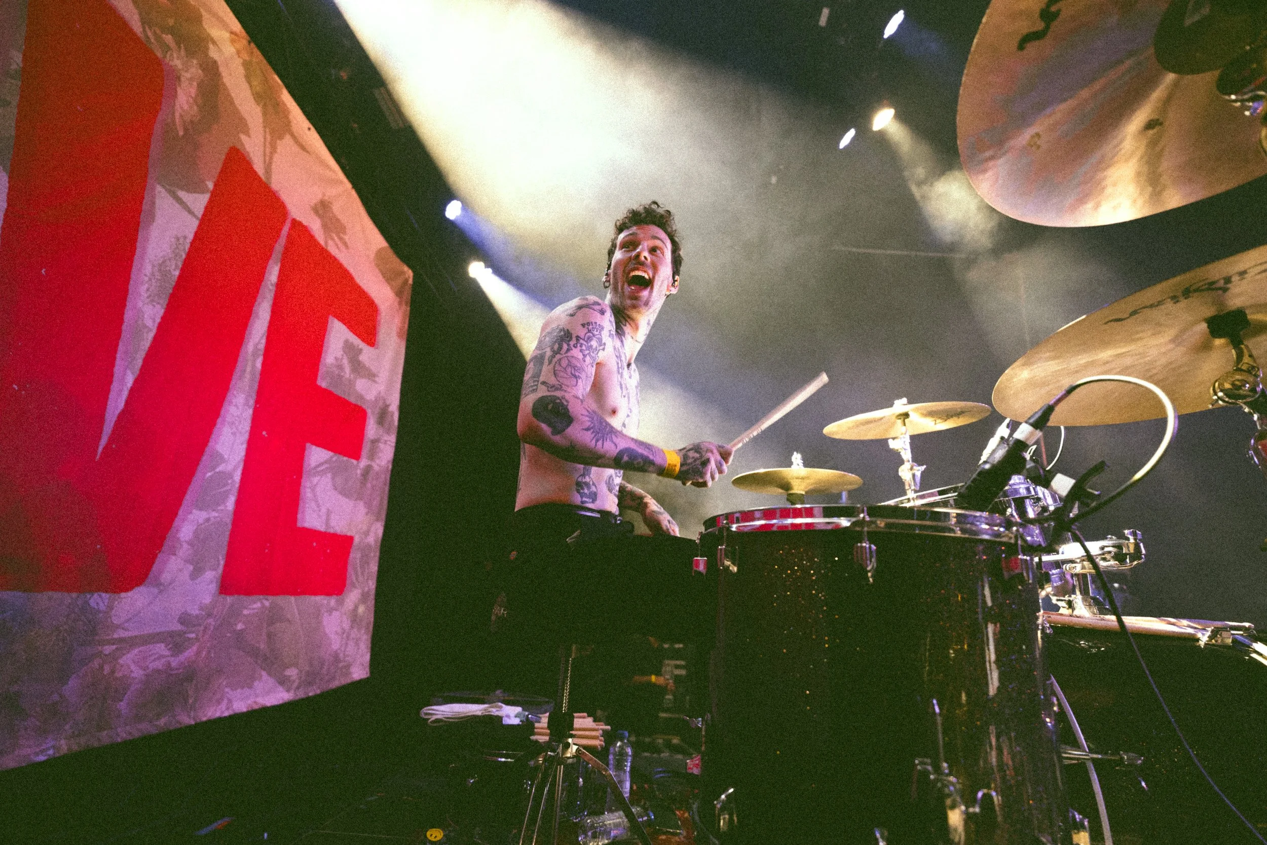 A shirtless drummer with tattoos is playing on stage, smiling with an expressive face. There is stage lighting and a large banner with red lettering behind him.