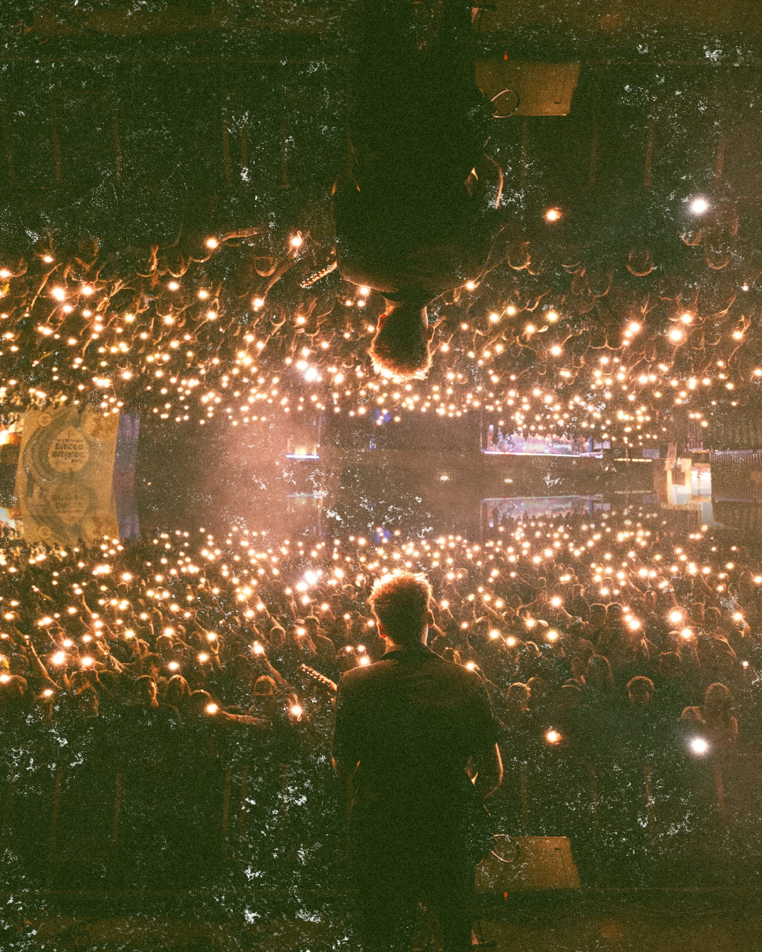A performer on stage facing an audience in a dimly lit venue with string lights overhead, and a large crowd holding up lights or phones creating a glowing effect.