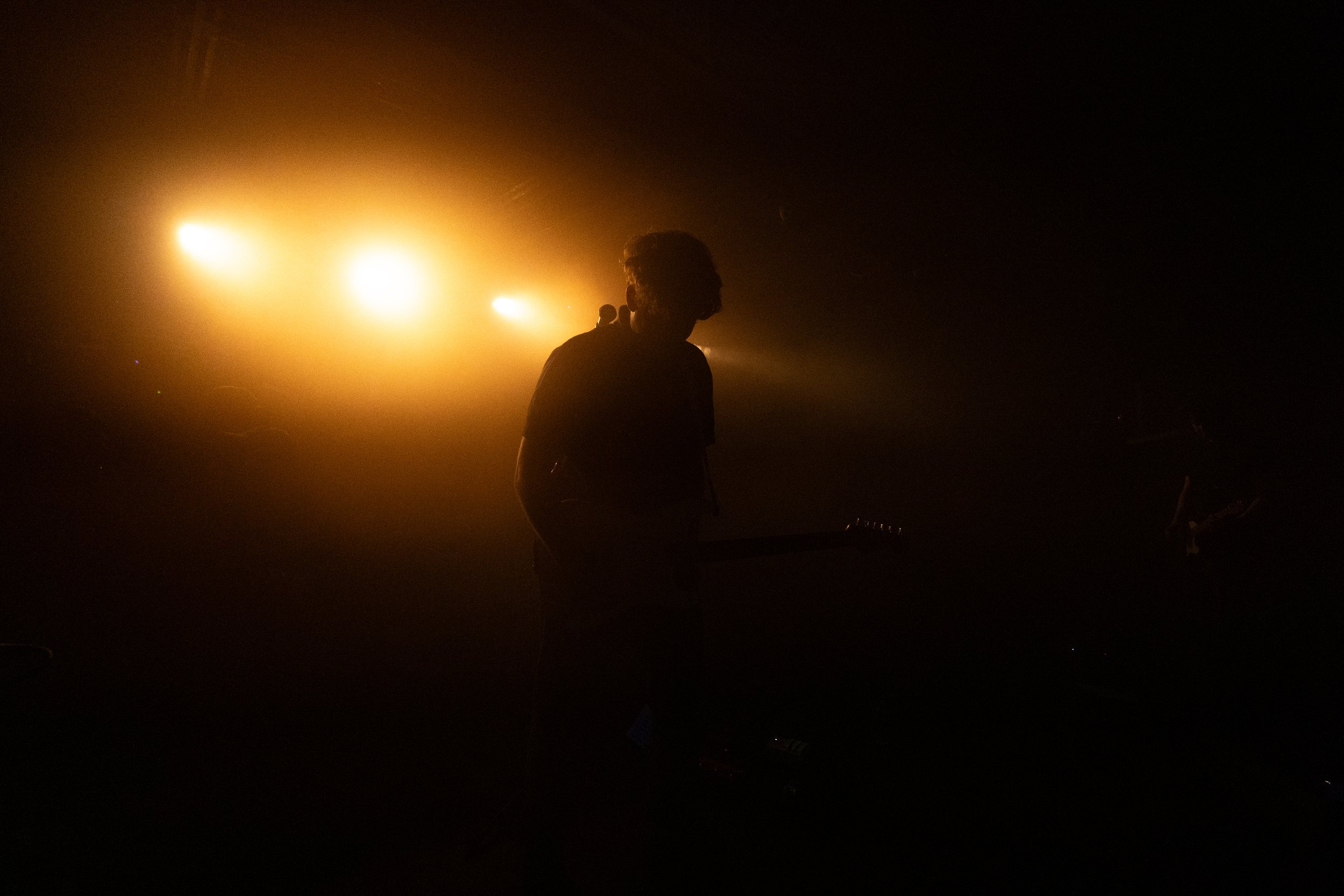 A person holding a guitar playing on stage with bright yellow-orange lights shining in the background.