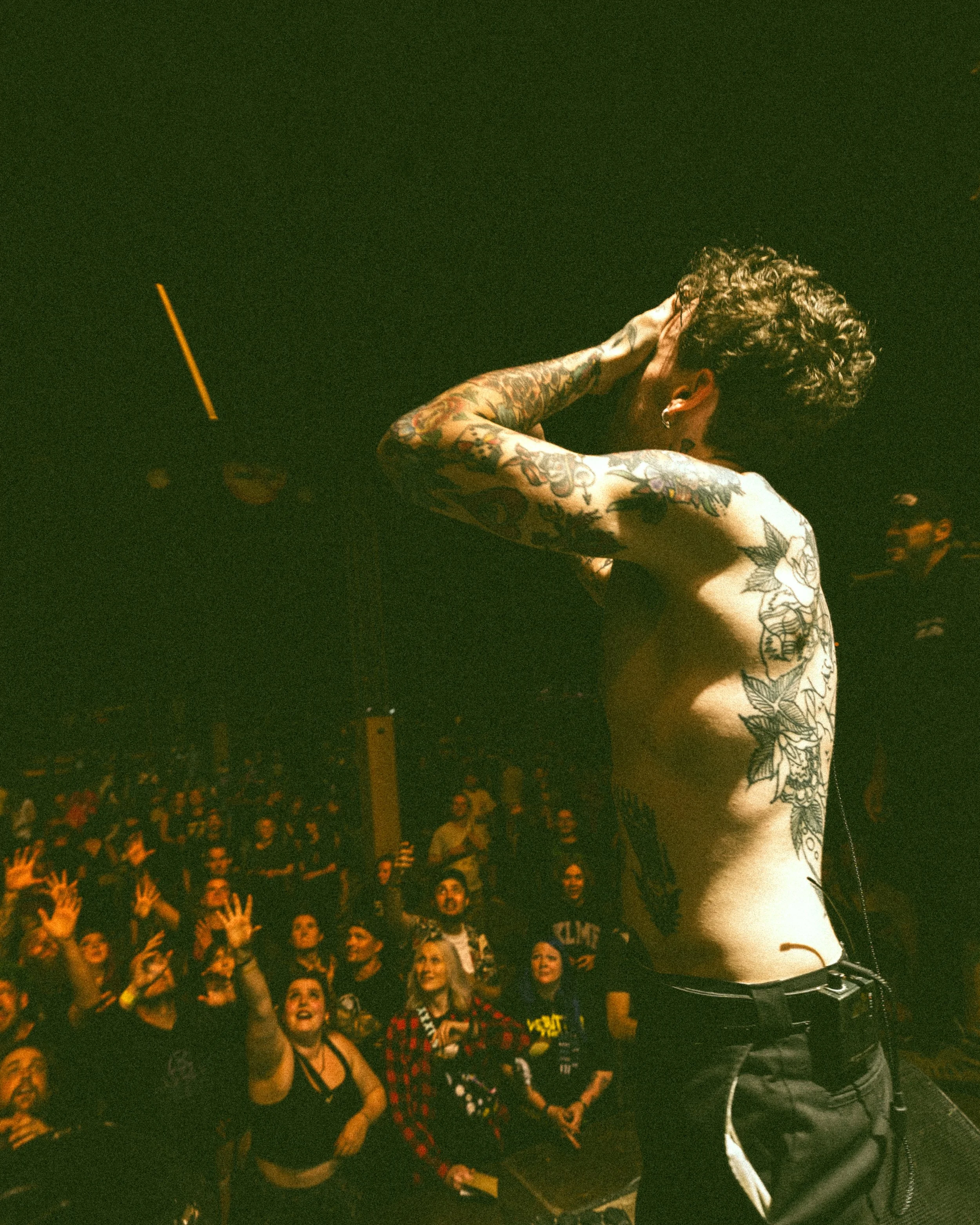 A shirtless person with tattoos performing on stage at a concert, facing sideways with their right hand behind their head. The crowd in the background is watching and reaching towards the performer.