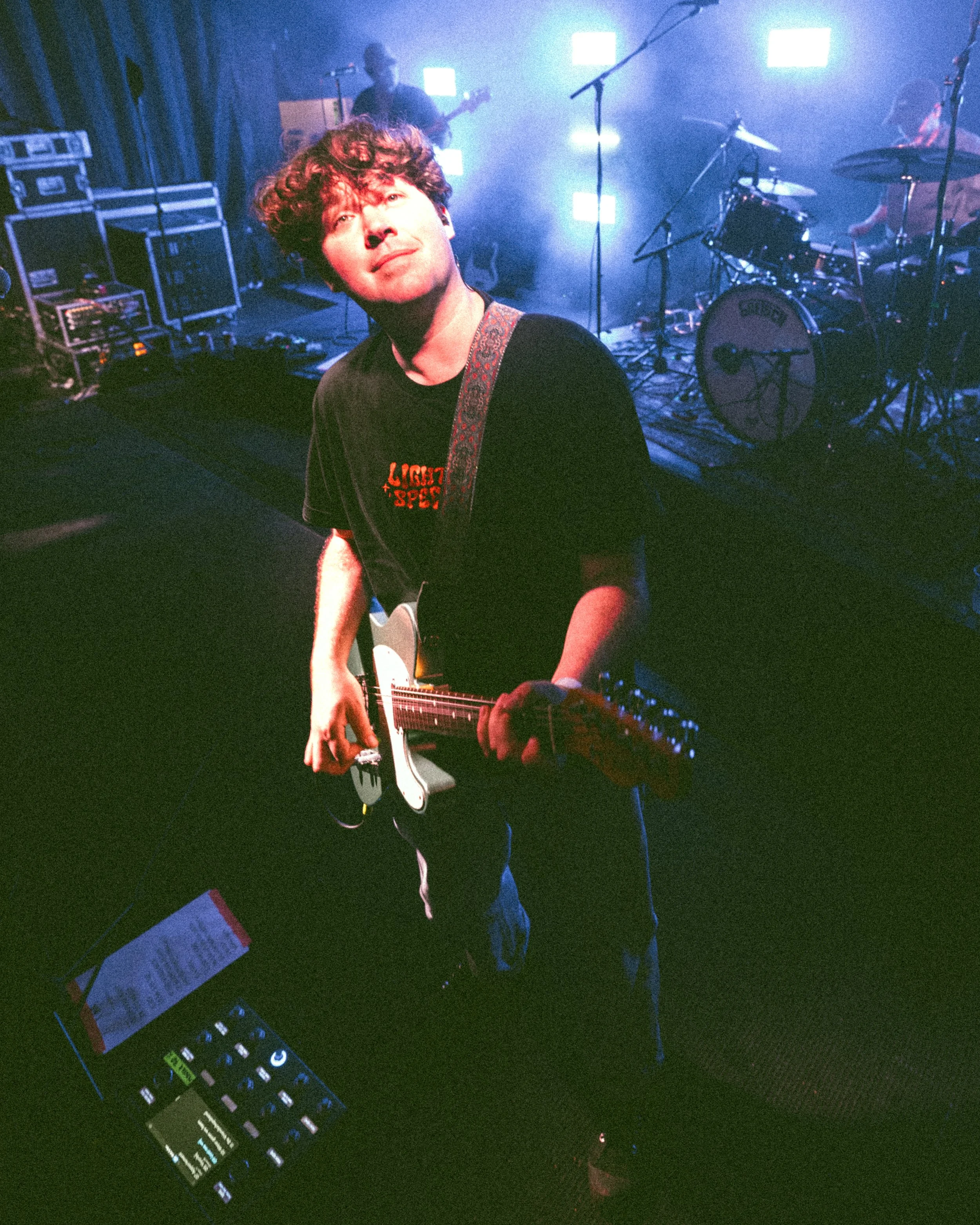 A young man with curly hair holding an electric guitar on stage with a band, with a drummer and a bassist in the background, stage lights create a vibrant atmosphere.