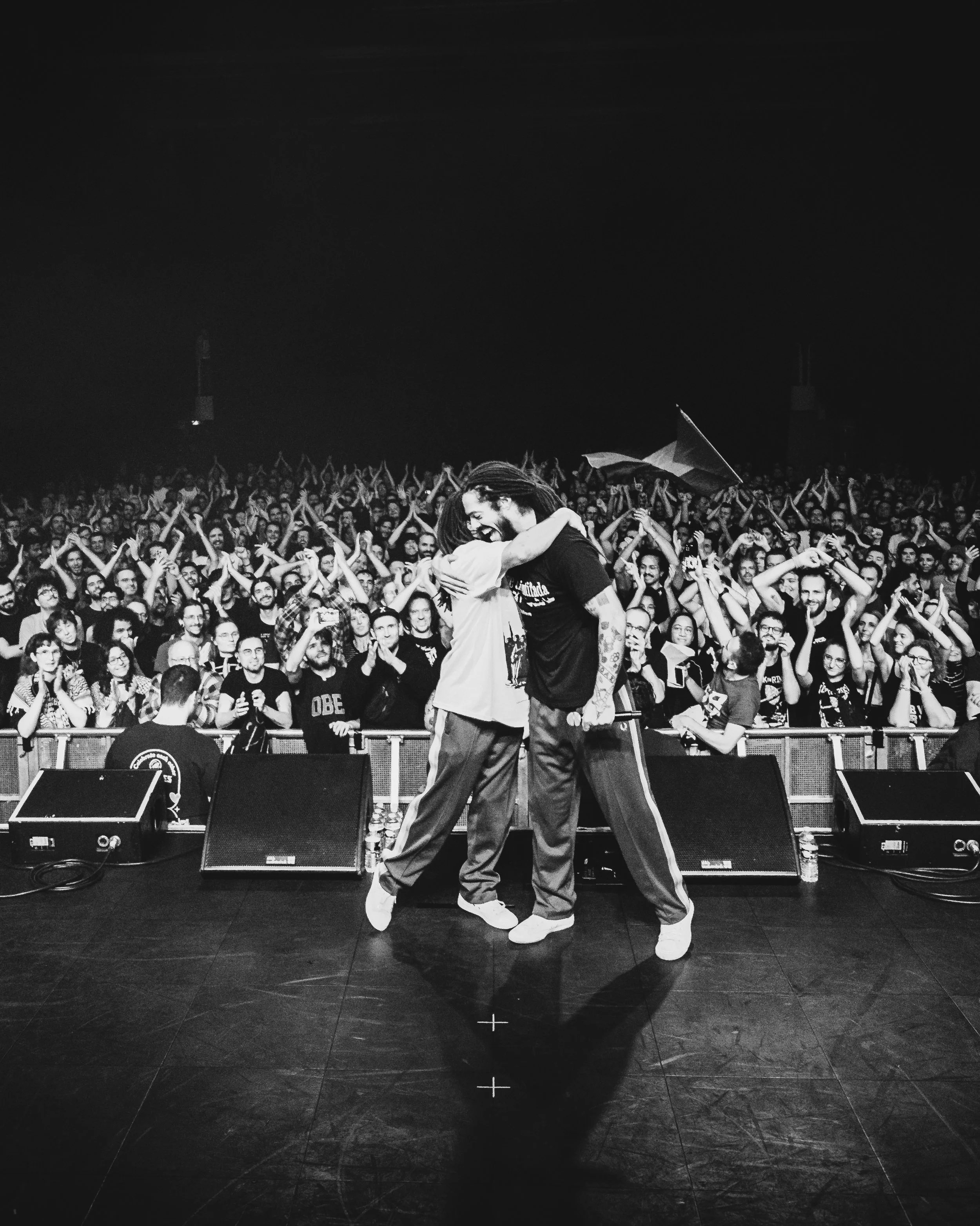Two performers hugging on stage in front of a large cheering crowd in black and white.
