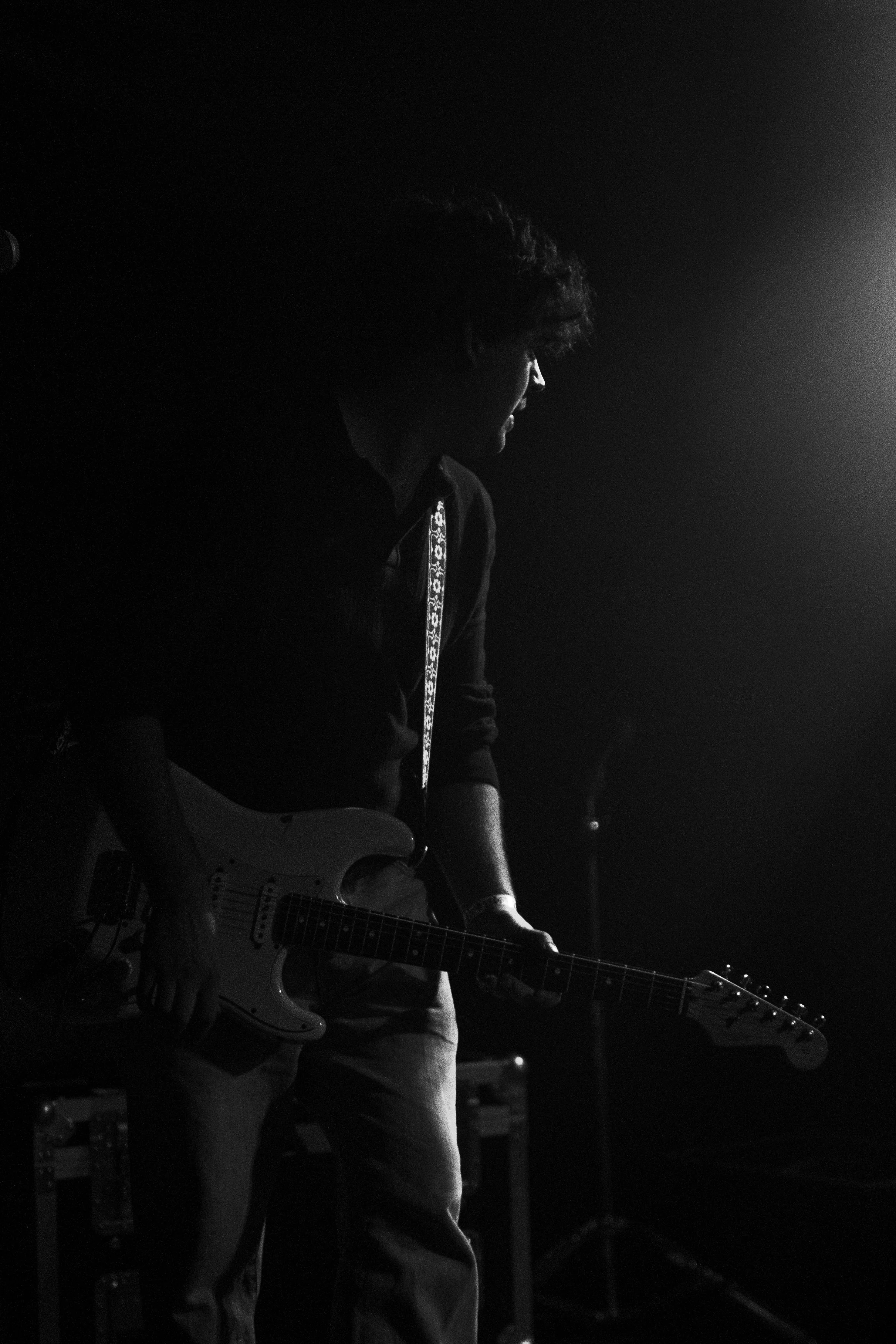 Silhouette of a person playing electric guitar on a dark stage with a bright light source behind them.