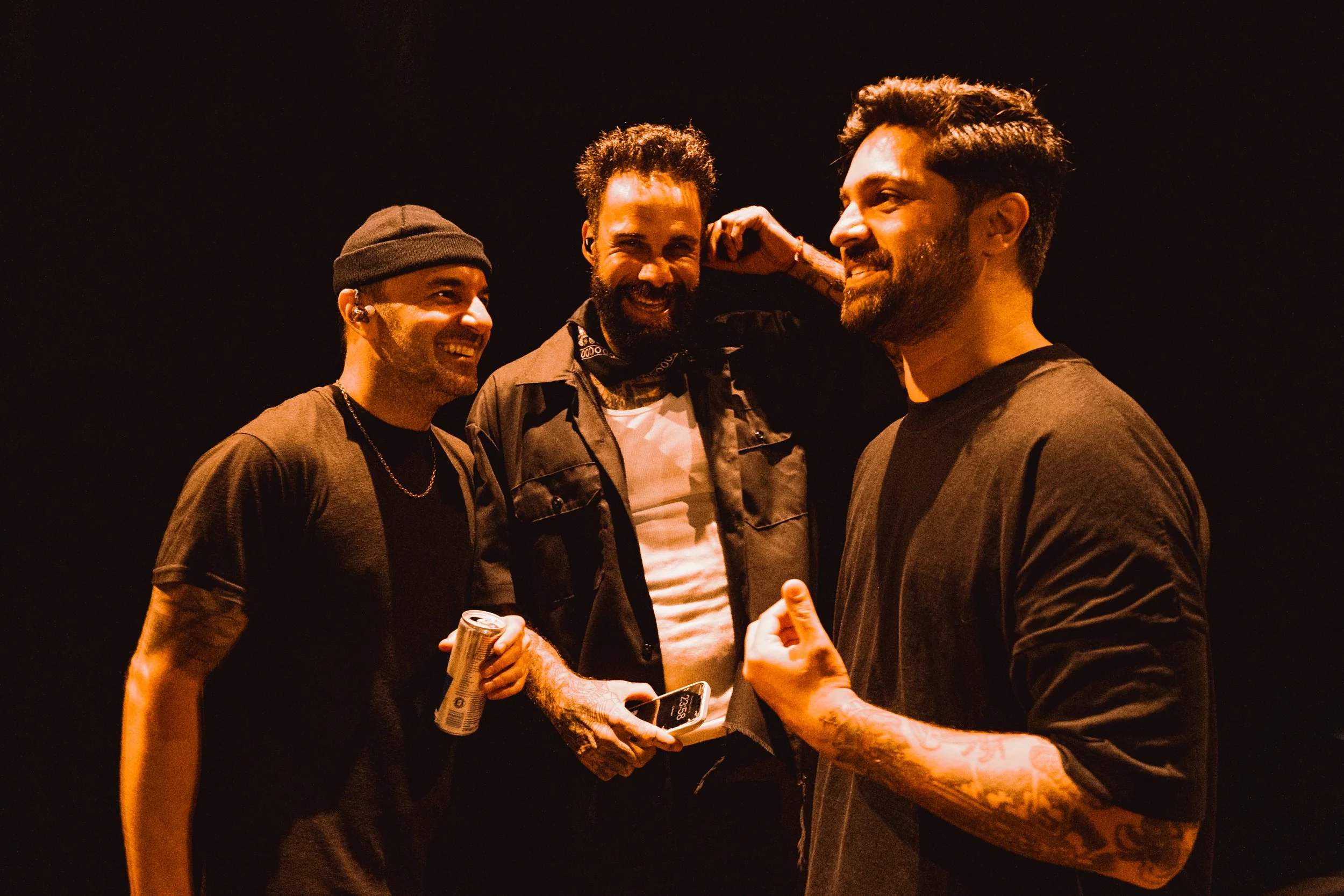 Three men in black shirts standing together, smiling, in a dark setting with warm lighting, one holding a soda can and another with tattoos on his arm, engaging in conversation and physical gestures.
